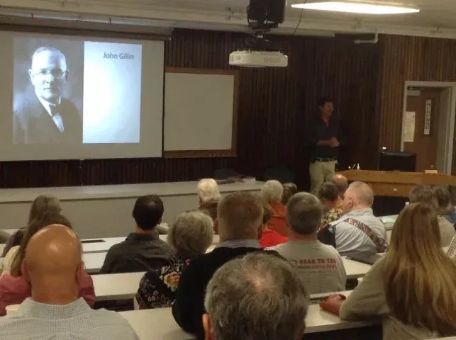 A man lectures in a classroom with a projector showing a portrait. Audience listens, seated at desks.
