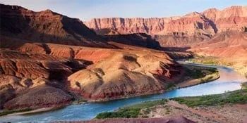 River flowing through a canyon with red rock formations. Blue water and a sunny sky are visible.