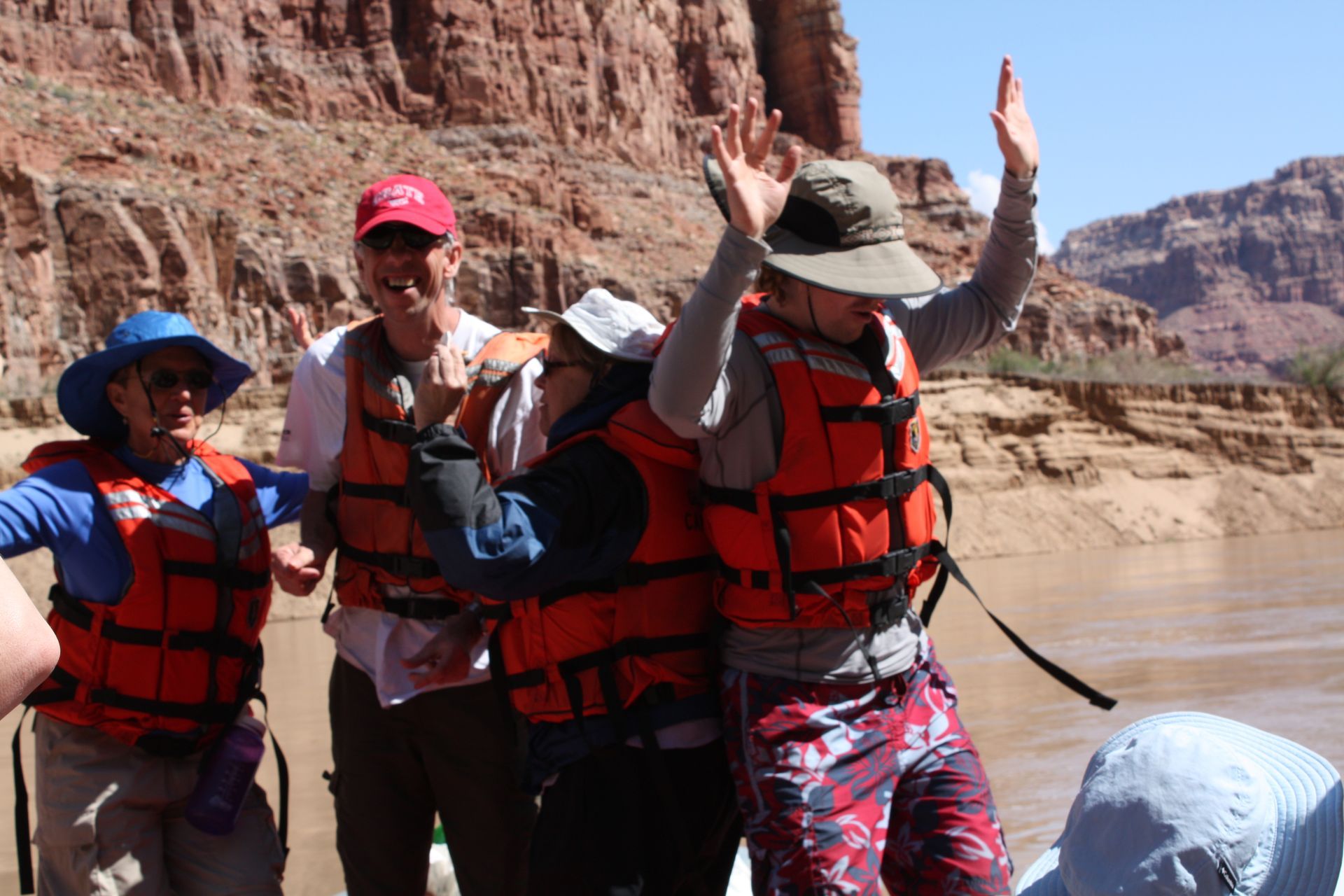 People on a raft in a canyon, wearing life jackets. One person has their arms raised.