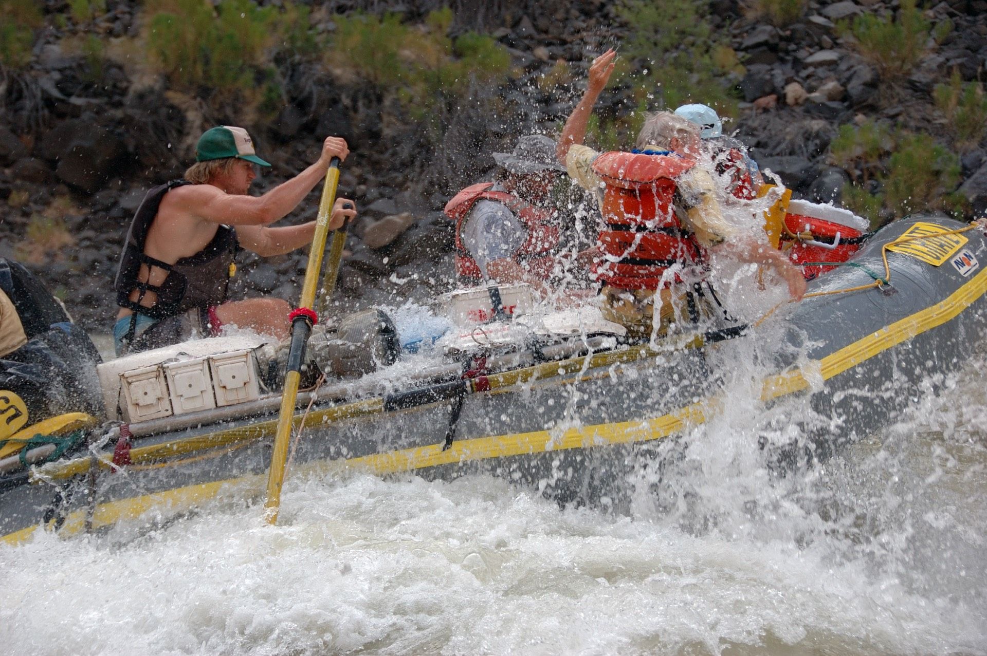 Two people on a raft in a canyon river, wearing life vests and hats.