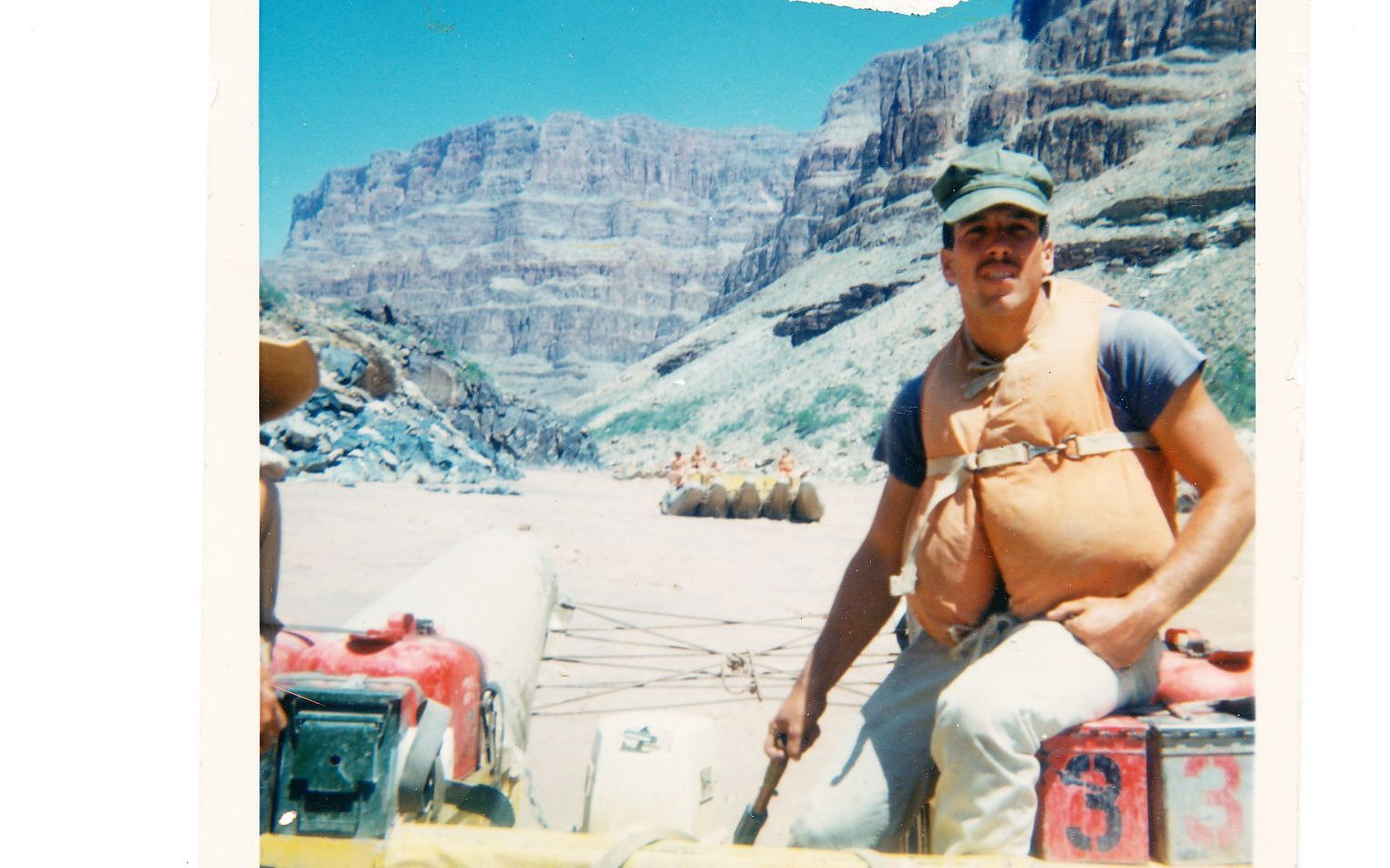 Man in life vest on raft in a canyon river, blue sky.