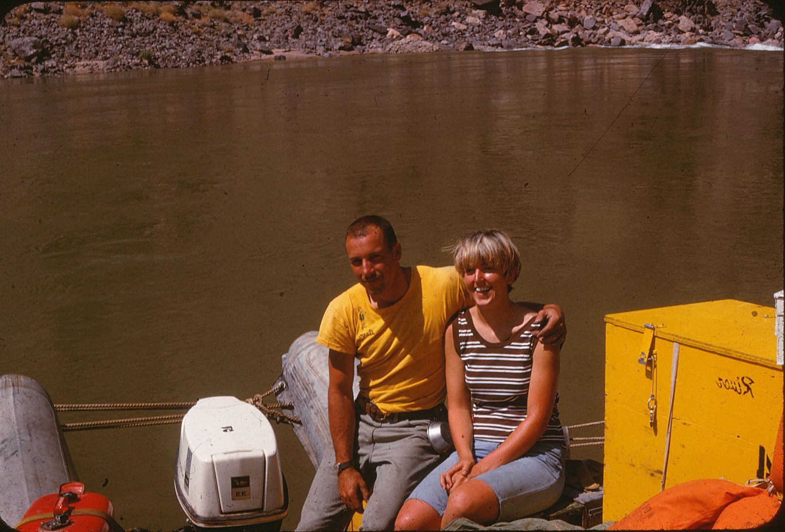 Couple on a boat on a river. Man in yellow shirt has arm around woman in striped top, both smiling.