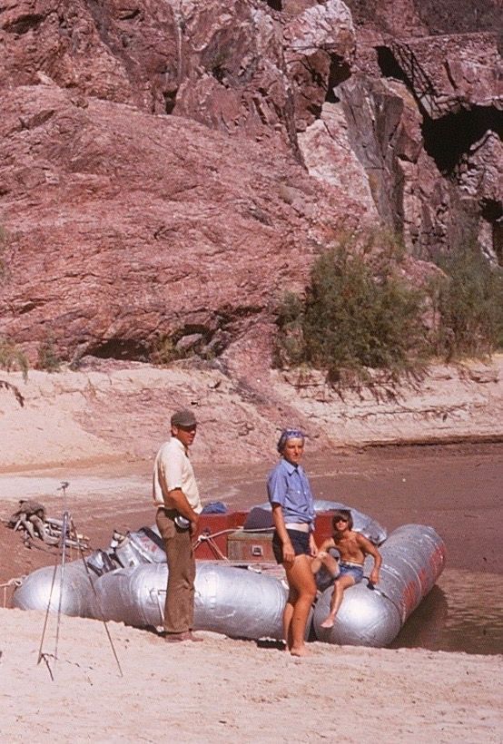 People by a raft on a sandy shore next to reddish rock formations. One person sits on the raft.