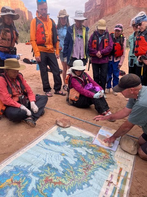 A group on a sandy riverbank, looking at a large map. One person points, others wear life vests and hats.