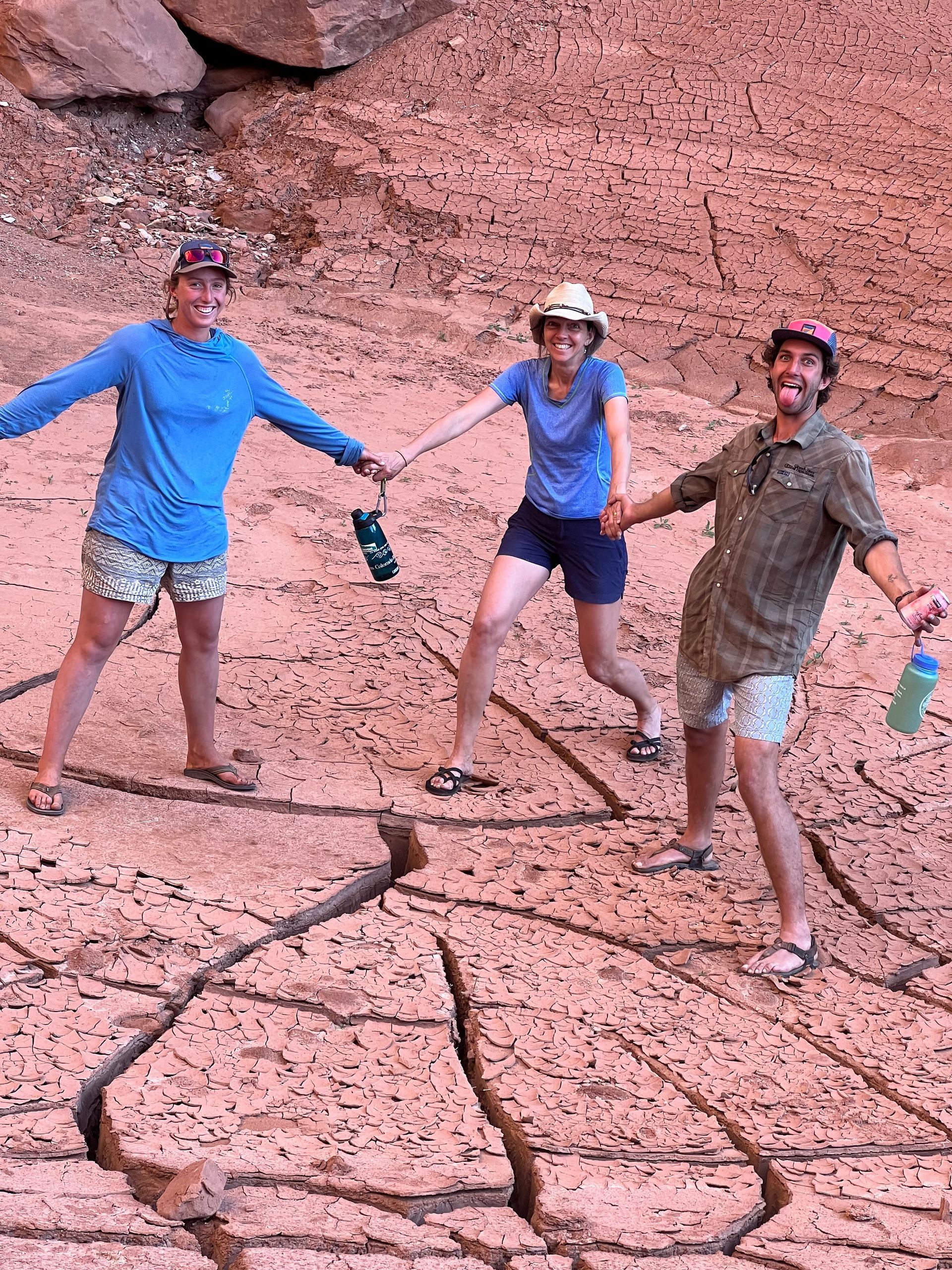 Three people holding hands on cracked, dry earth; one in blue, one in tan, one in green, all smiling.