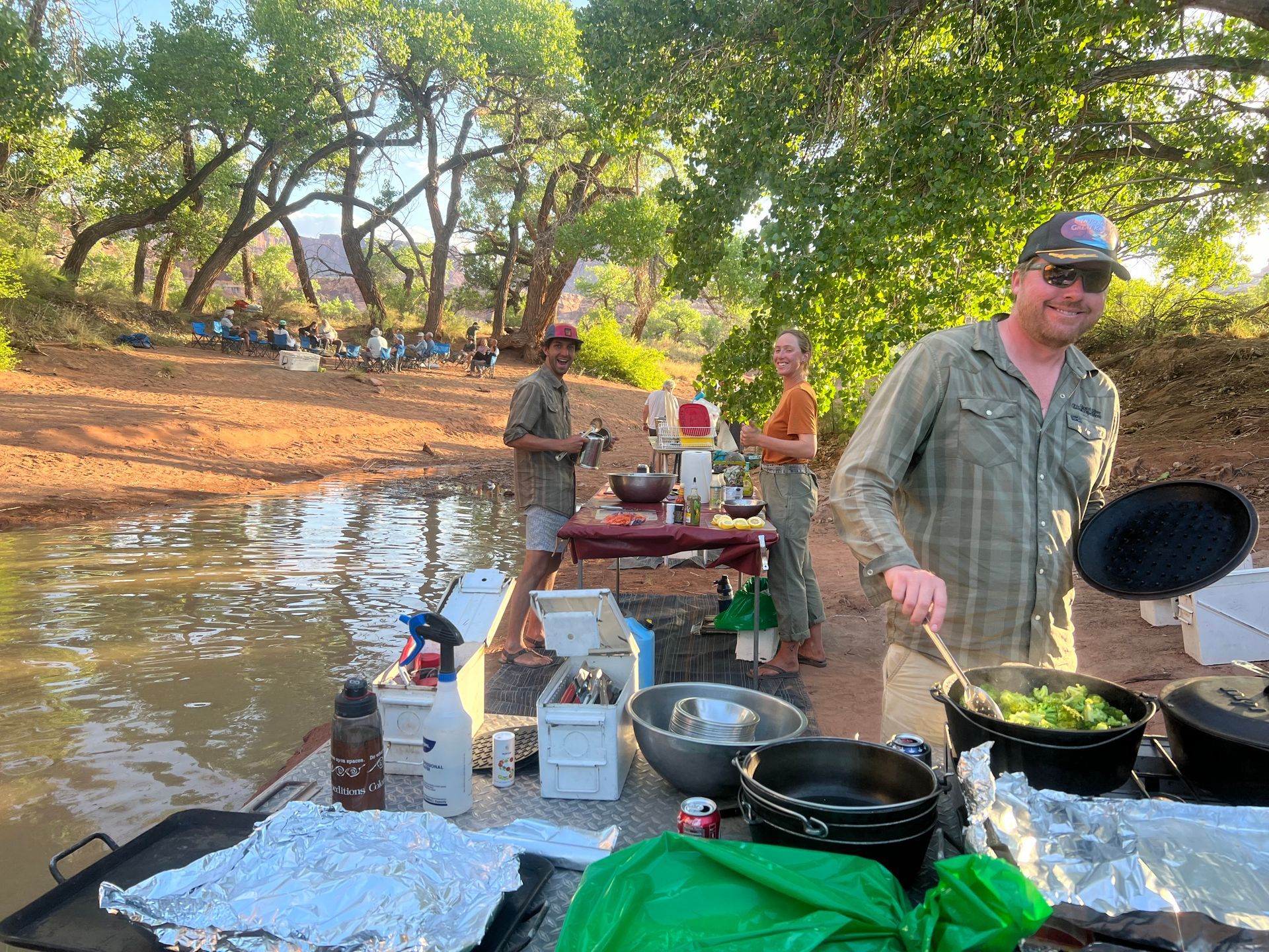 People cooking and eating outdoors near water. Trees and red earth in the background.