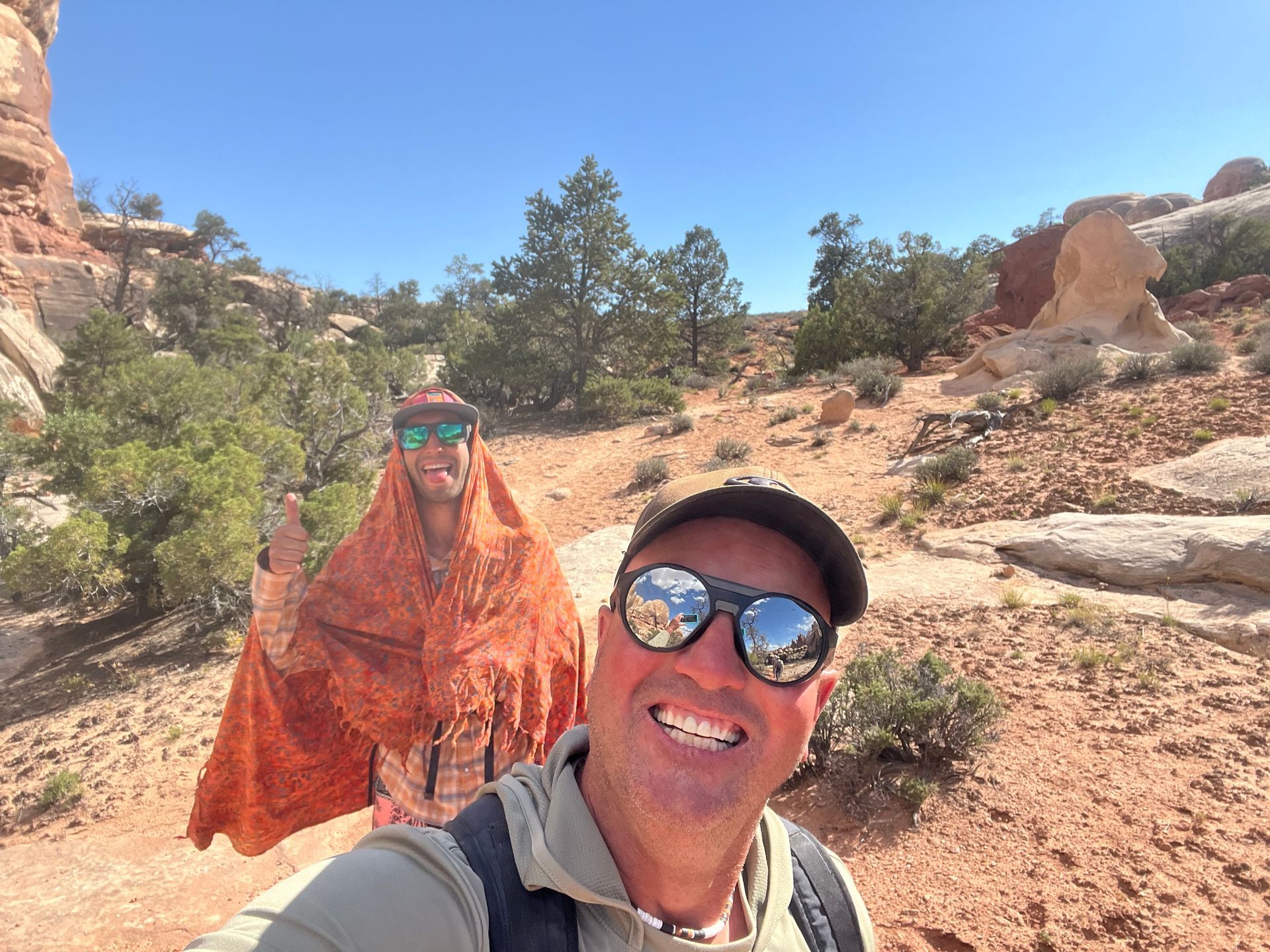 Two people hiking on a red rock trail under a blue sky; one smiles at the camera, the other wears an orange shawl.
