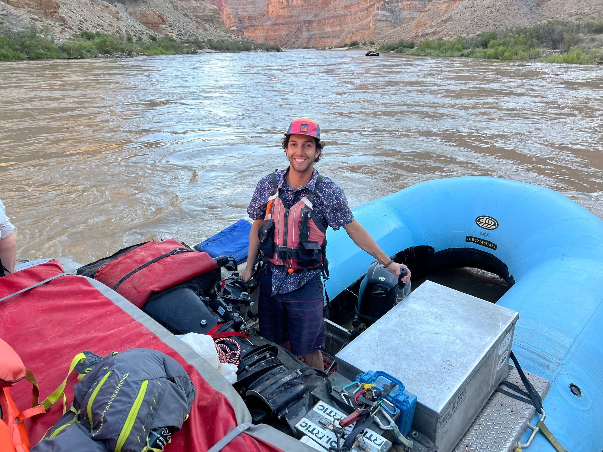 Man in life vest smiles on a raft on a muddy river, surrounded by gear and canyon walls.