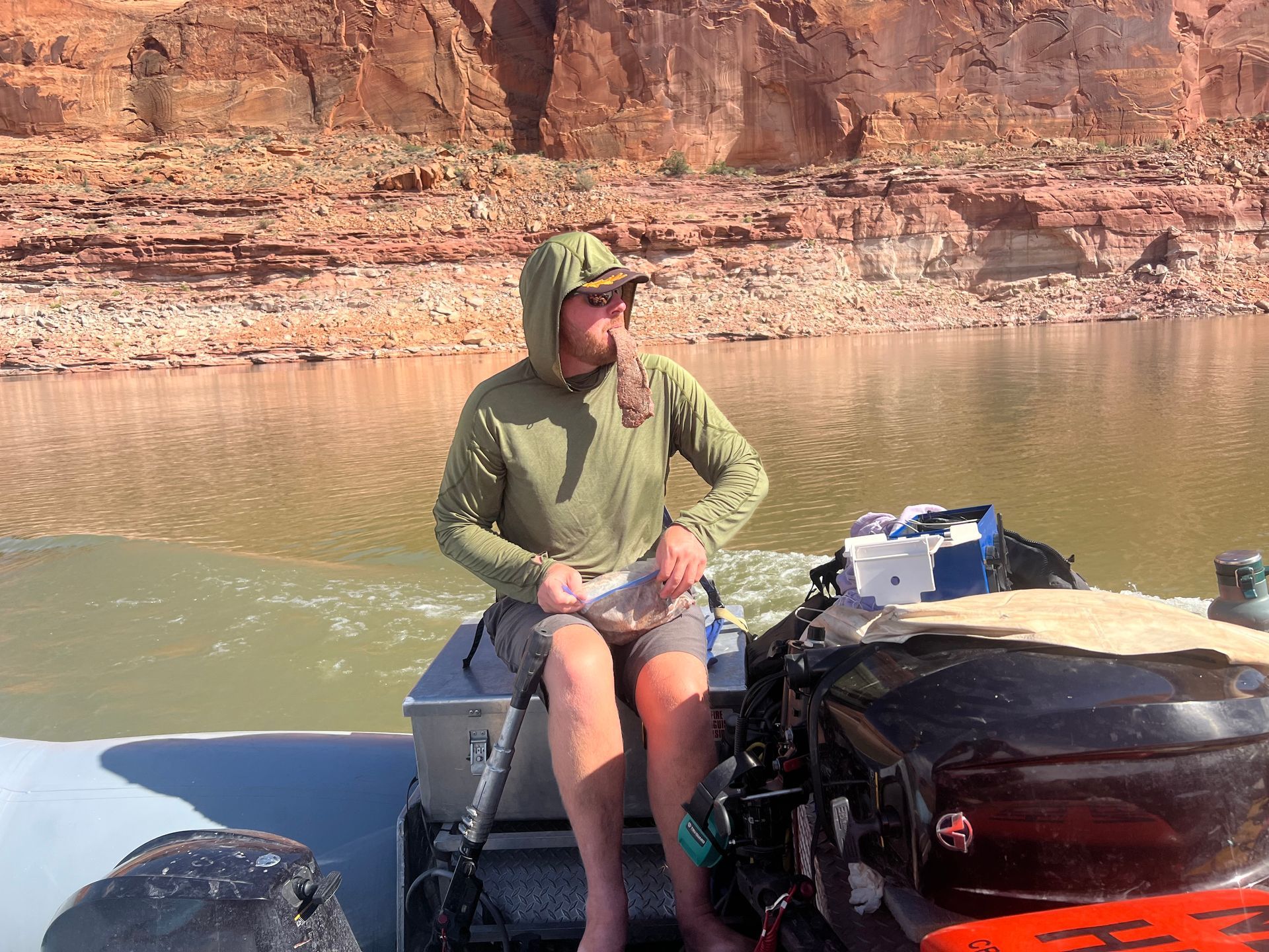 Man in green hooded shirt on a motorboat, looking at something off-screen, with red rock canyon in the background.