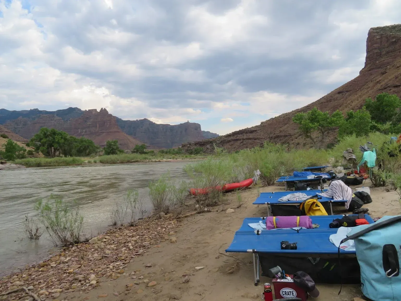 Camping setup on a sandy riverbank, with cots, gear, and mountains in the background. Cloudy sky.