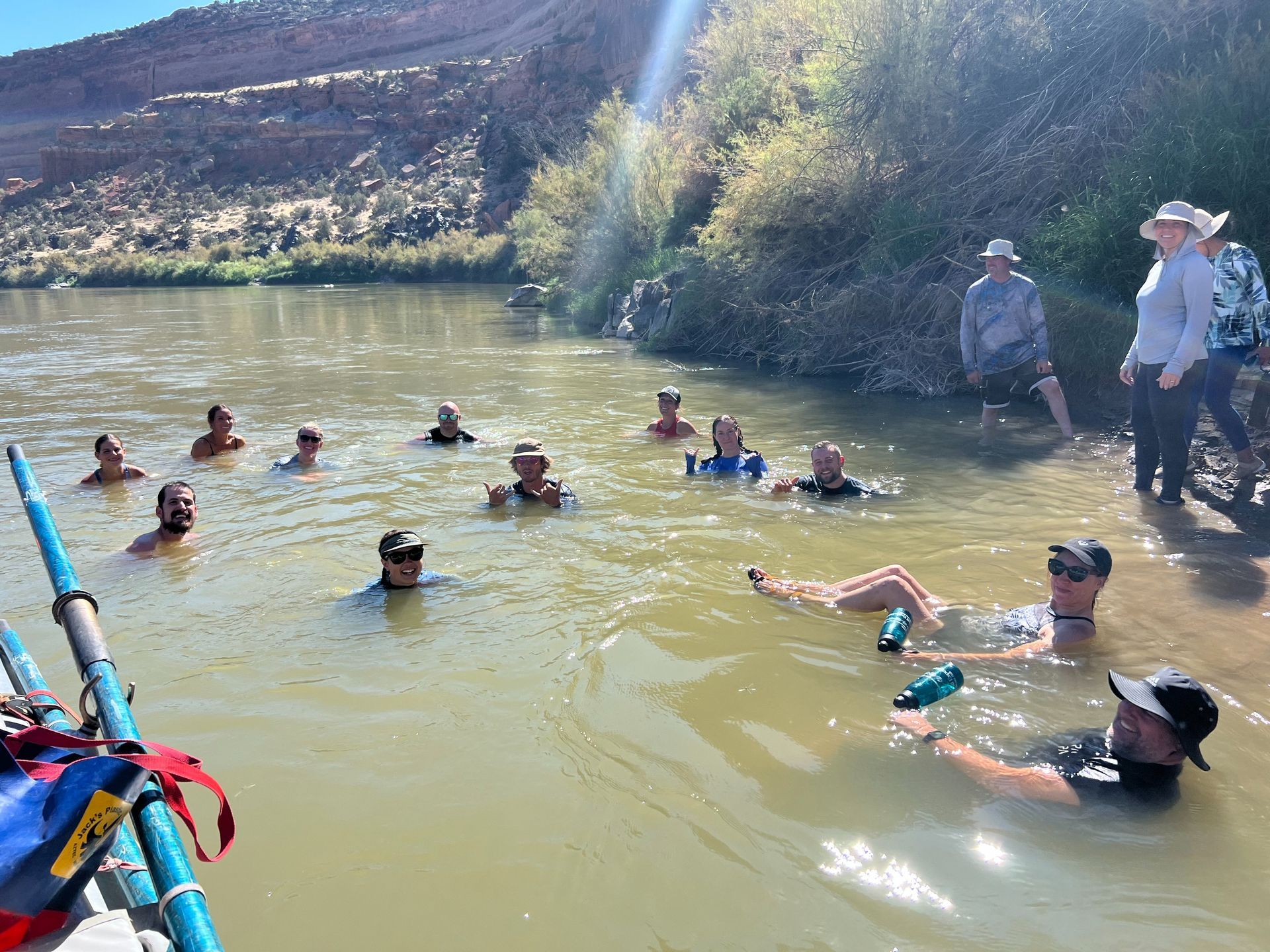 People swimming in a river next to a bank with trees and some standing people. Sunny day.