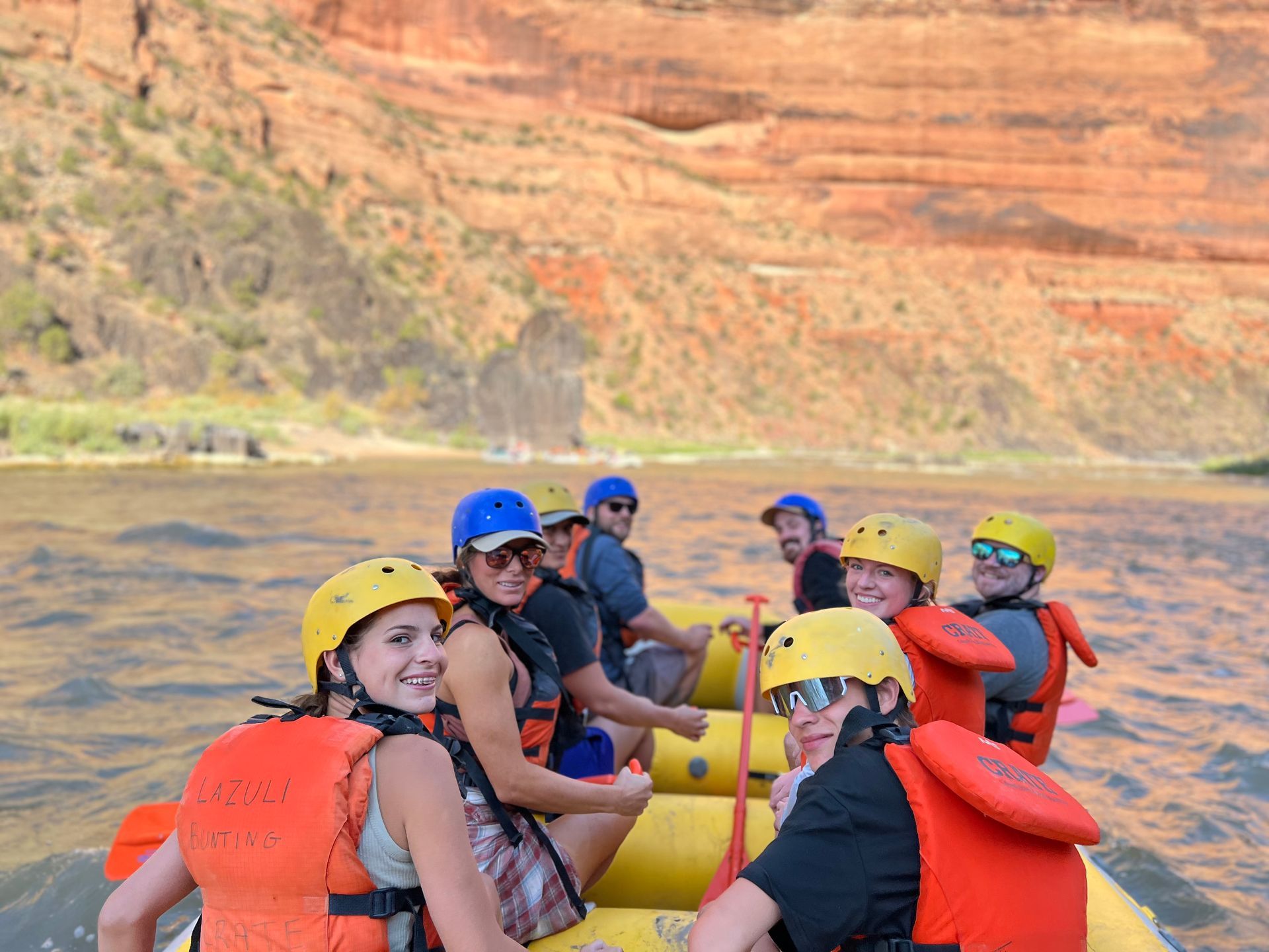 People on a yellow raft, wearing helmets and life vests, on a river with red rock cliffs in the background.