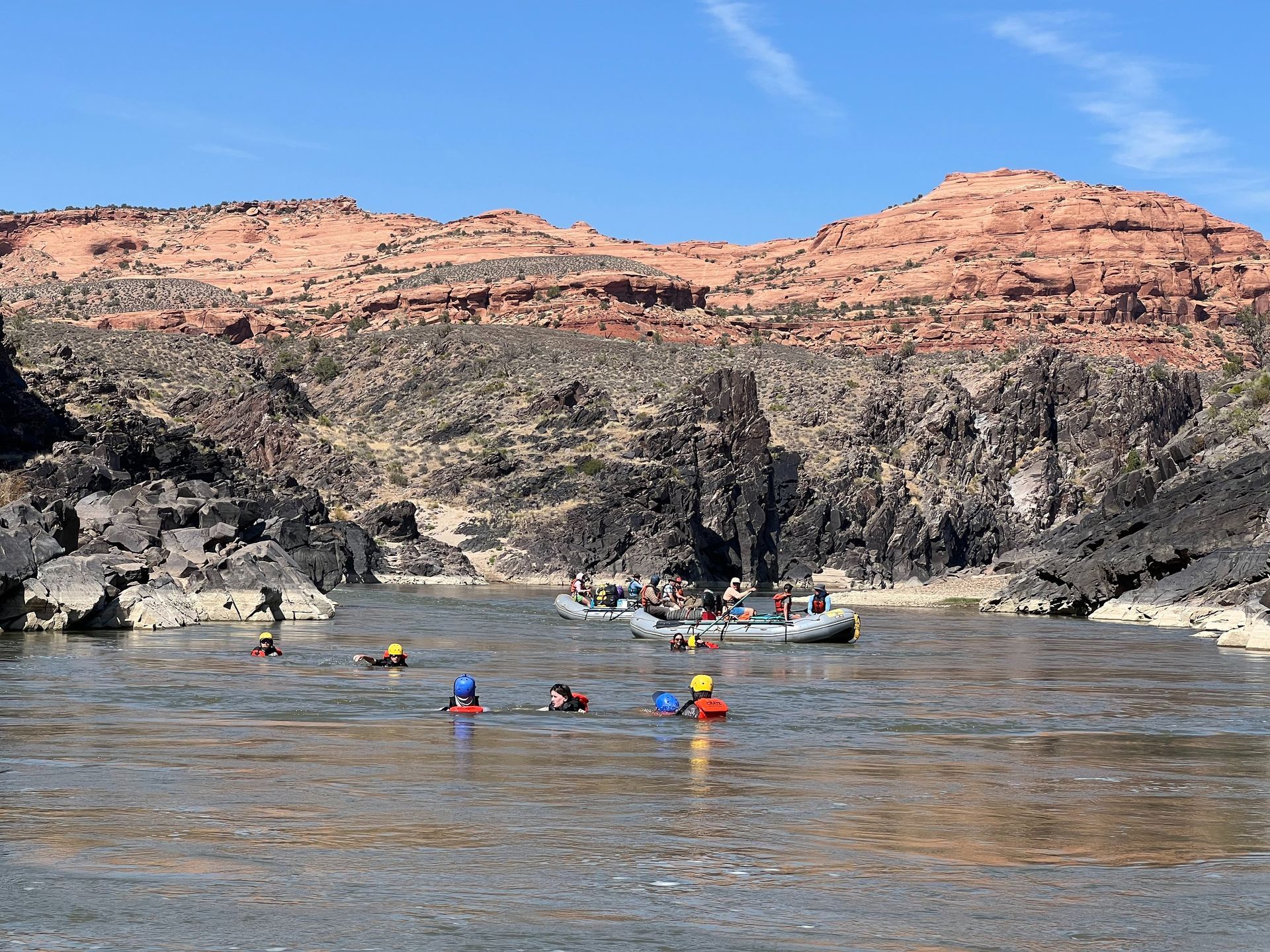 Rafters in a river near red and black rock formations under a blue sky.