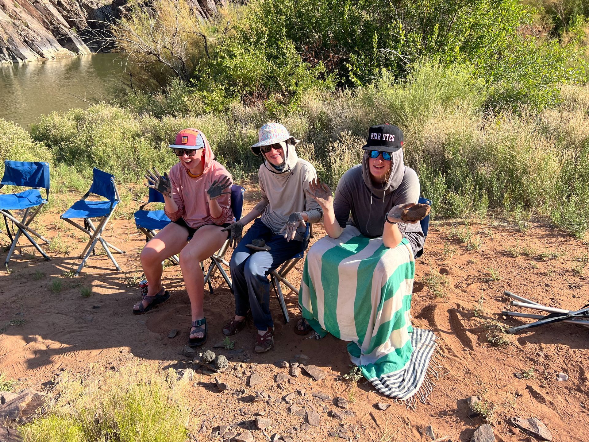 Three people sitting by a river, holding rocks. Green, blue, and white striped towel. Sunny day.