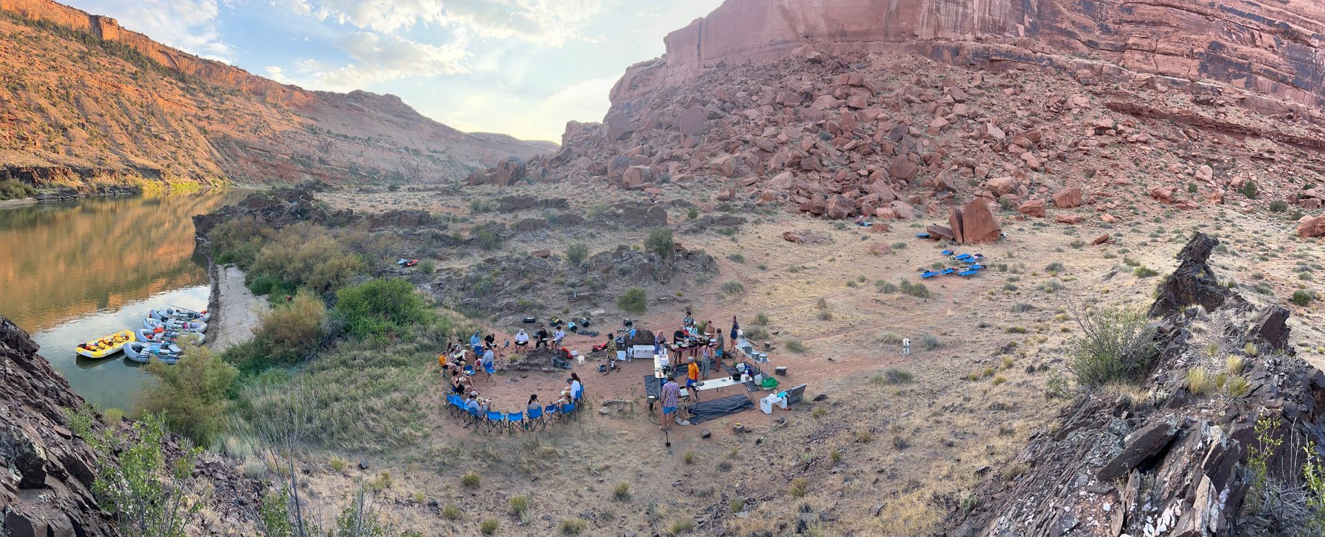 Panoramic view of a group of people gathered in a desert canyon. River and boats on the left, tents in the distance.