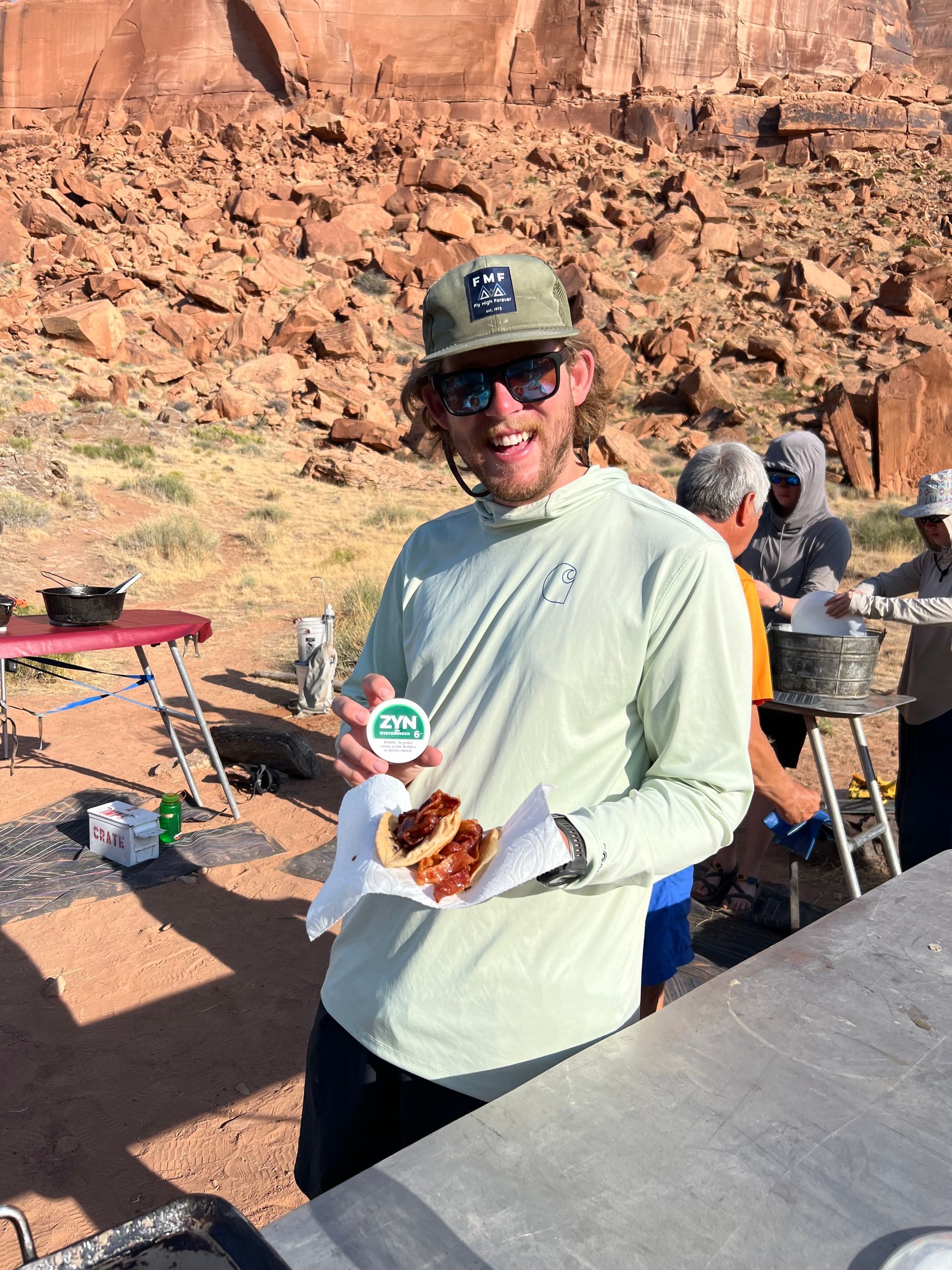 Man holding a sandwich, smiling, wearing sunglasses and hat, in a desert setting near a table with others.