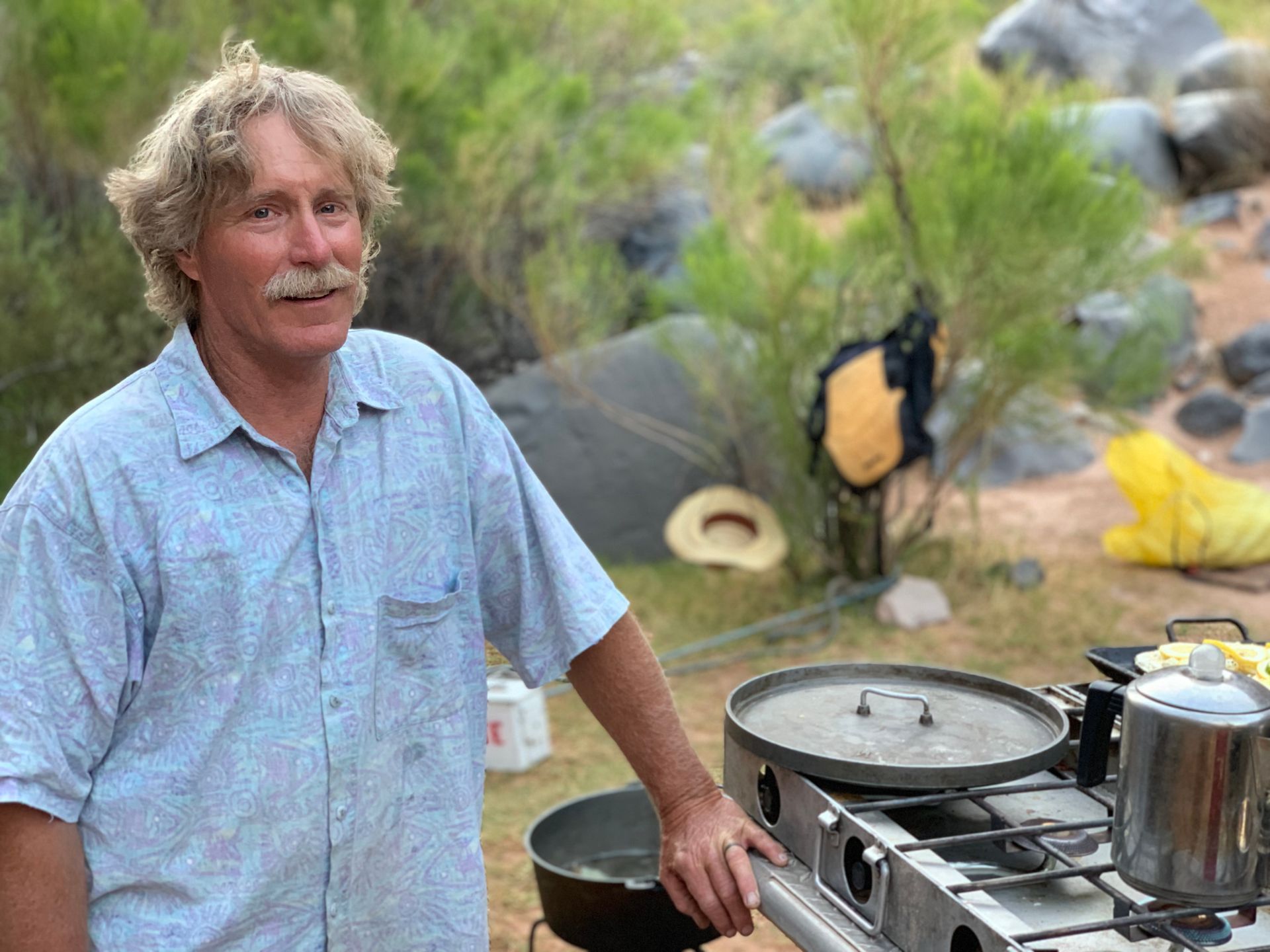 Man with curly hair and mustache by camp stove, cooking outdoors.