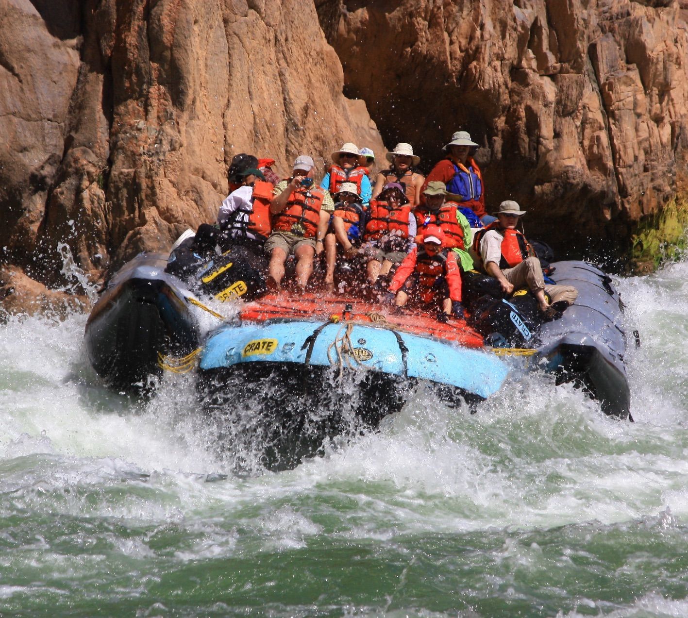 People rafting down a white water river in a blue and black raft, splashing through rapids.