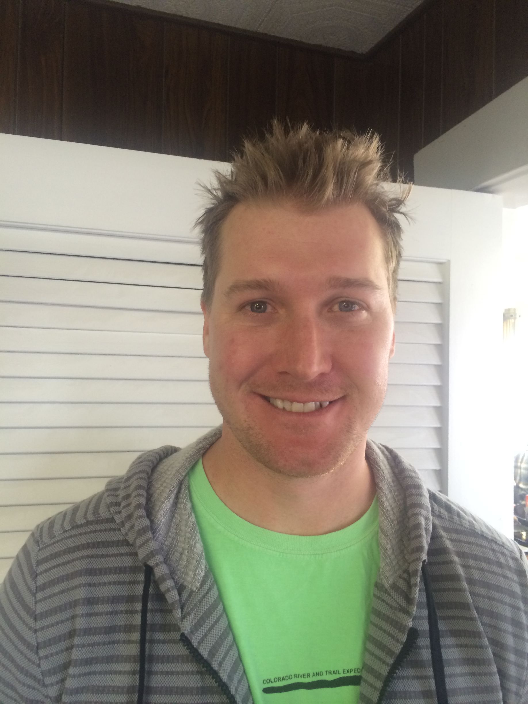 Man with spiky blond hair, wearing a gray hoodie and green shirt, smiling in front of a white wall.