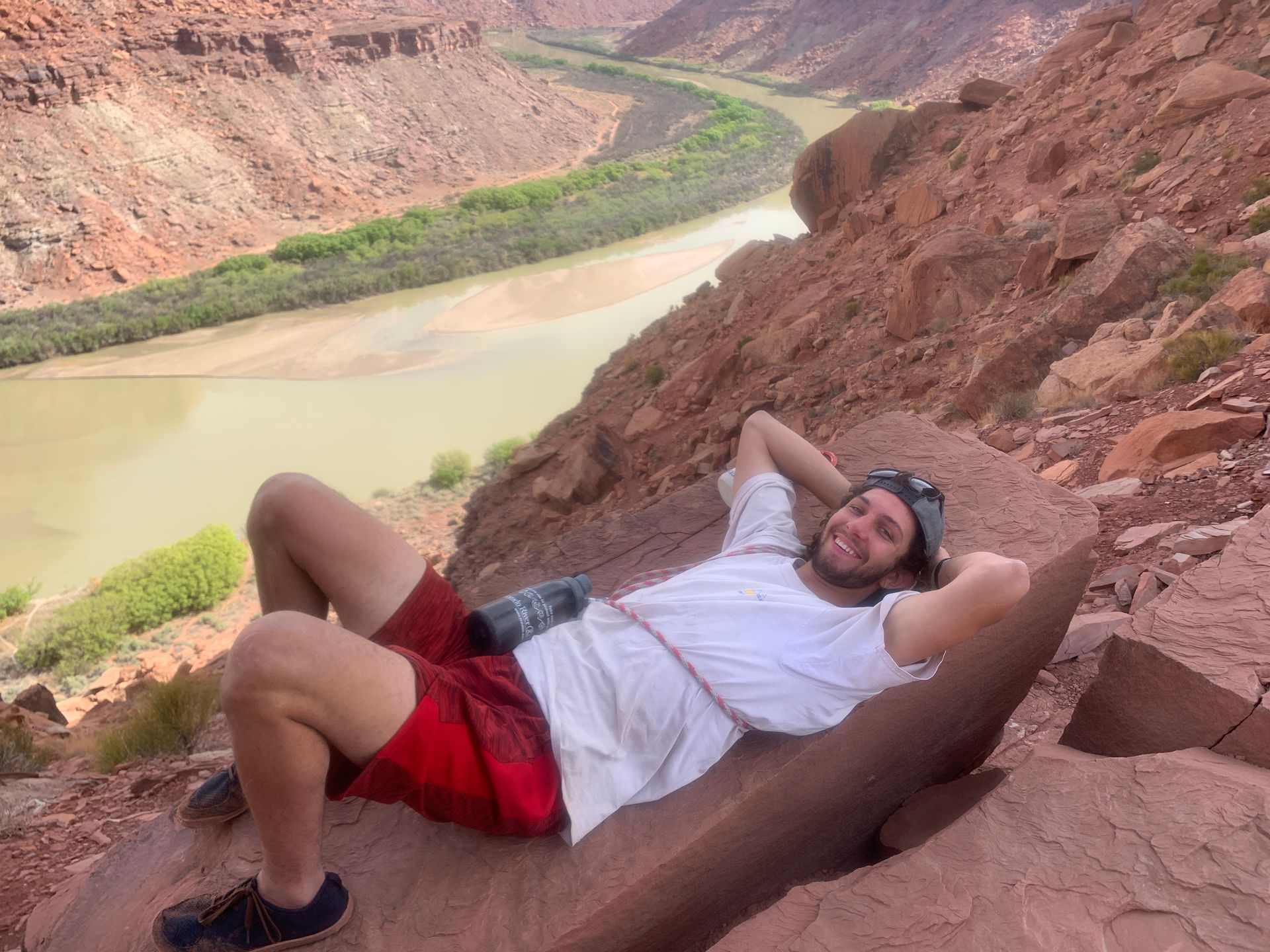 Man relaxing on a red rock hillside overlooking a river. He's wearing red shorts and a white shirt.