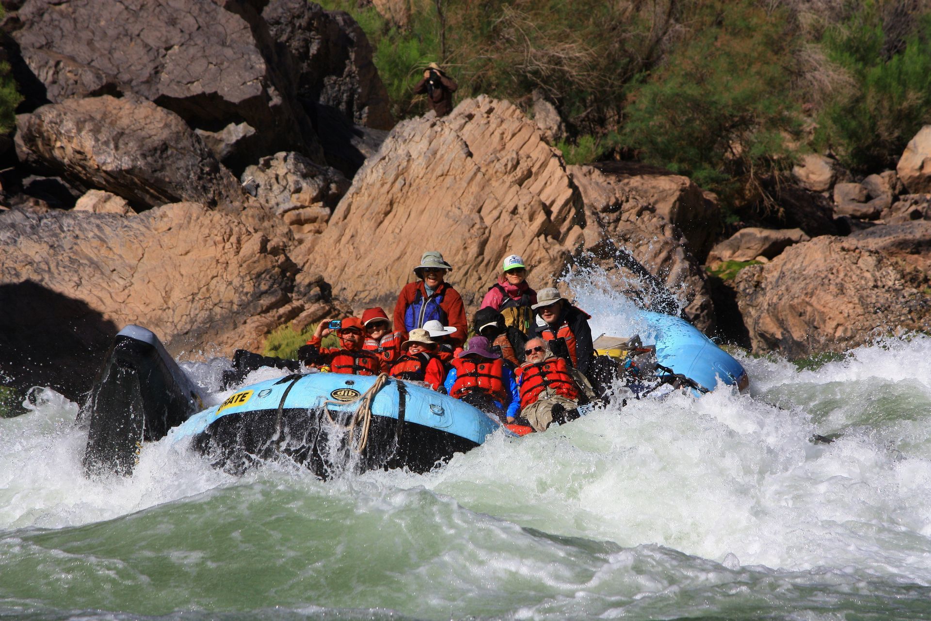 Rafting on a river with rapids; people wearing life vests, water splashing around, rocky setting.