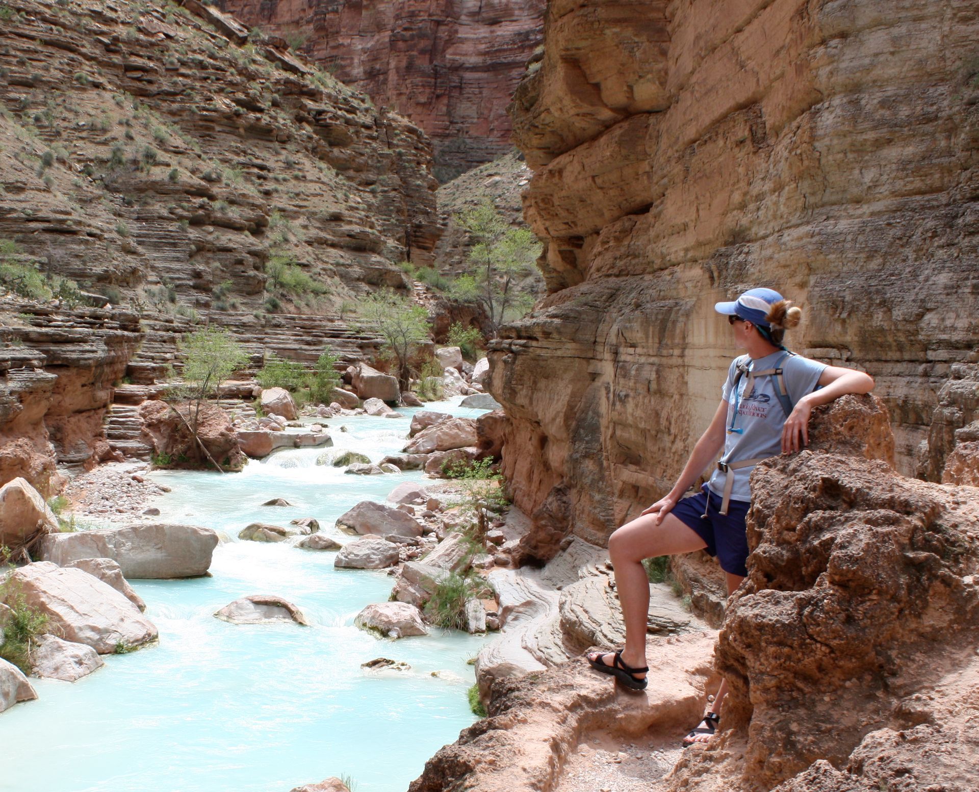 Woman sits on a cliffside, looking at a turquoise river in a canyon.