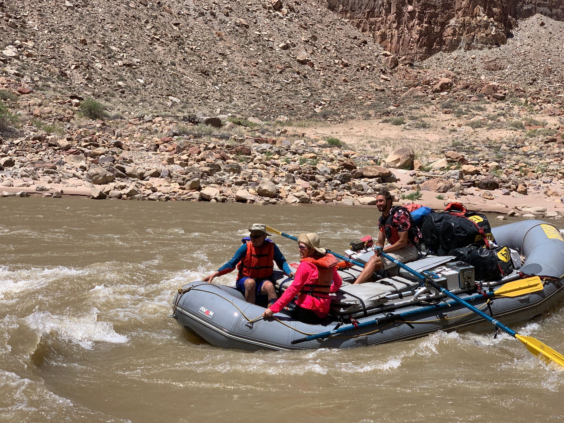 People on a raft navigate a river with rocky banks. The raft is grey; people wear life vests.