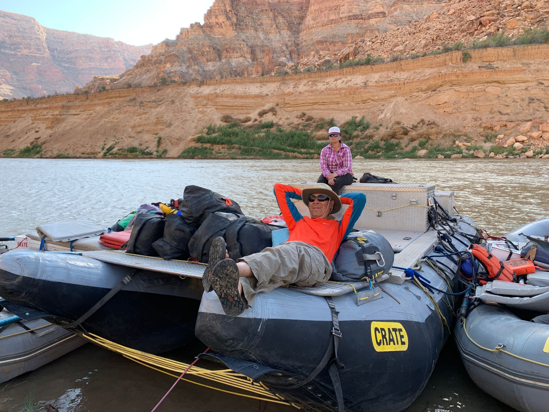 Person relaxing on a raft, hands behind head, other person in background near riverbank with desert landscape.