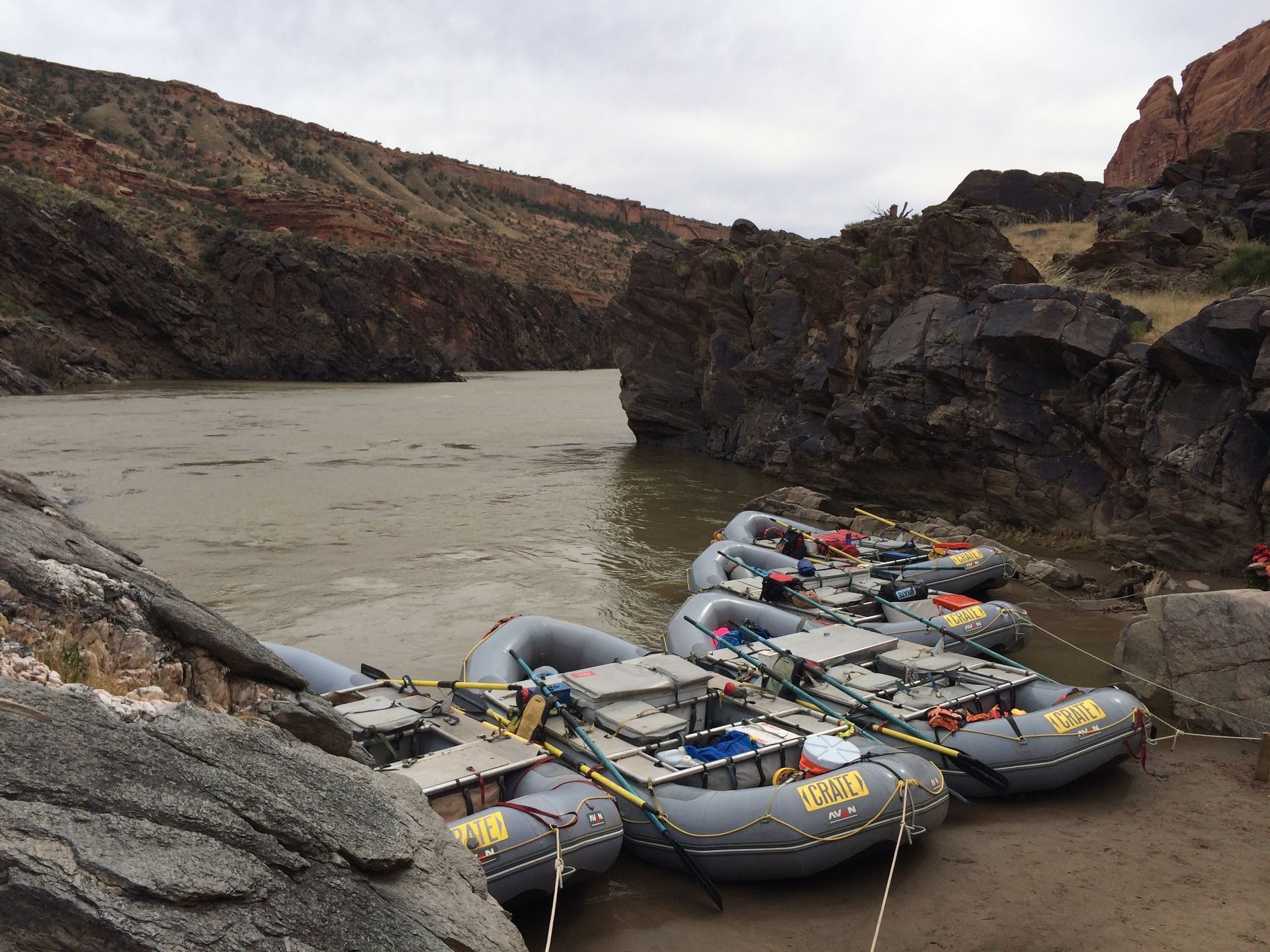 Rafts docked on a muddy riverbank. Brown water, rocky canyon walls, cloudy sky.