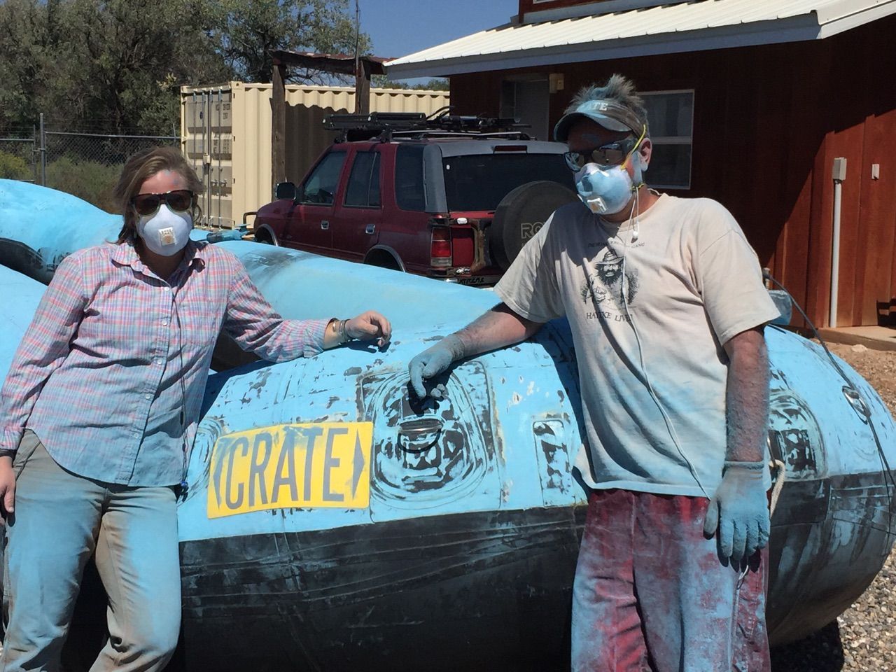 Two people in masks and paint-covered clothes next to a blue raft outdoors.