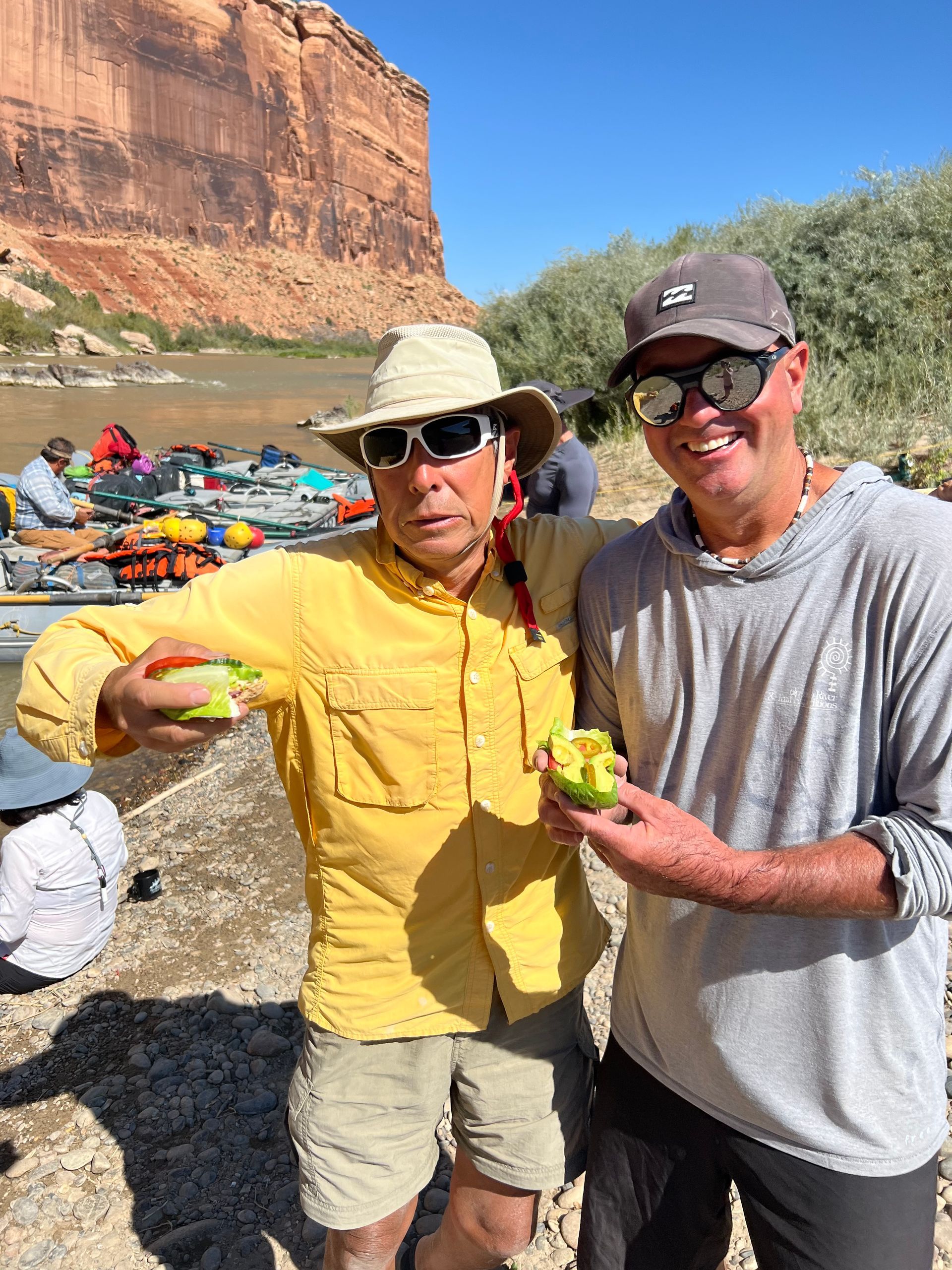 Two people hold yellow objects near a river. They wear hats and sunglasses, with boats and cliffs in the background.