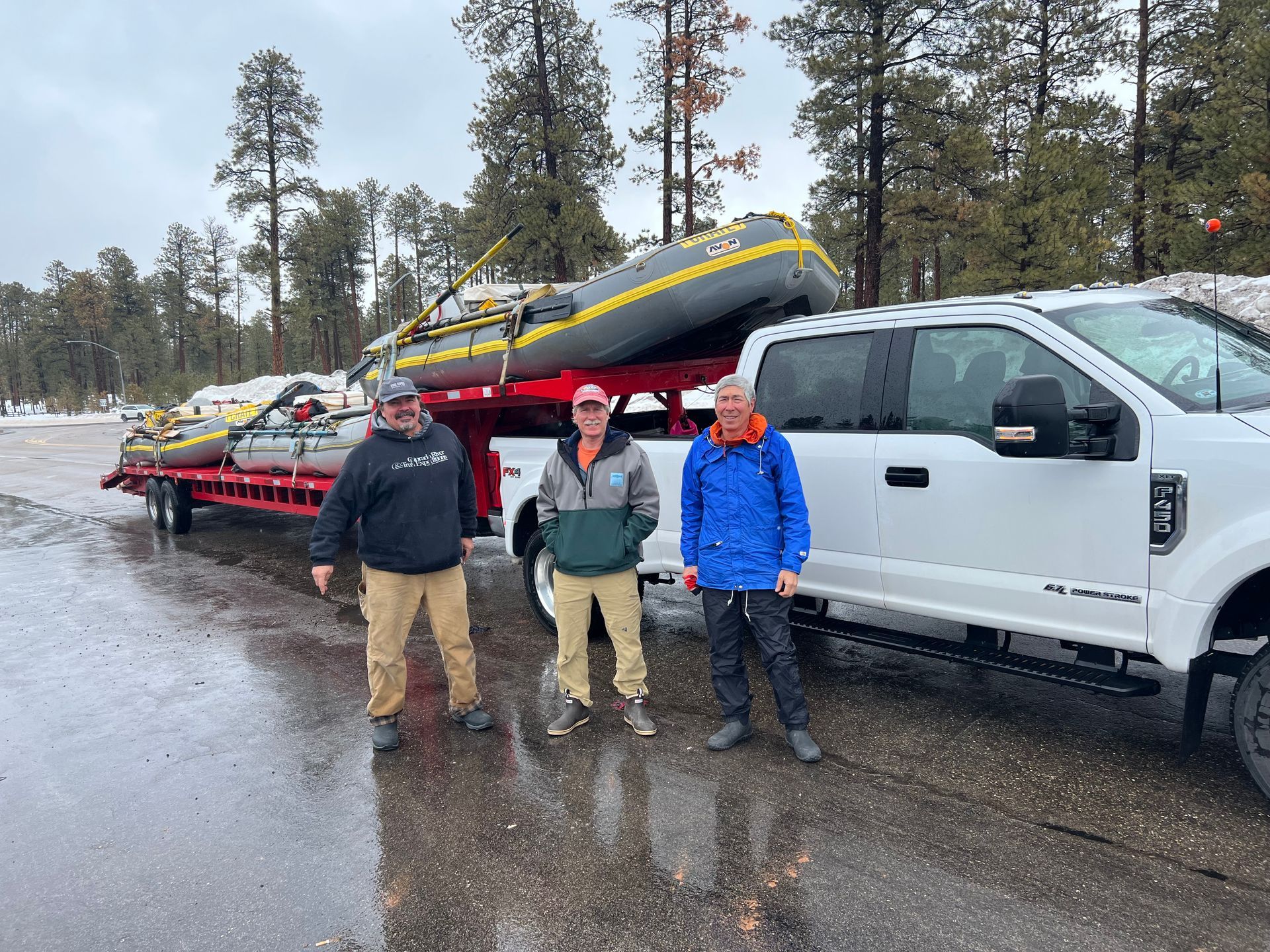 Three men stand next to a white truck towing a trailer with two inflatable rafts.
