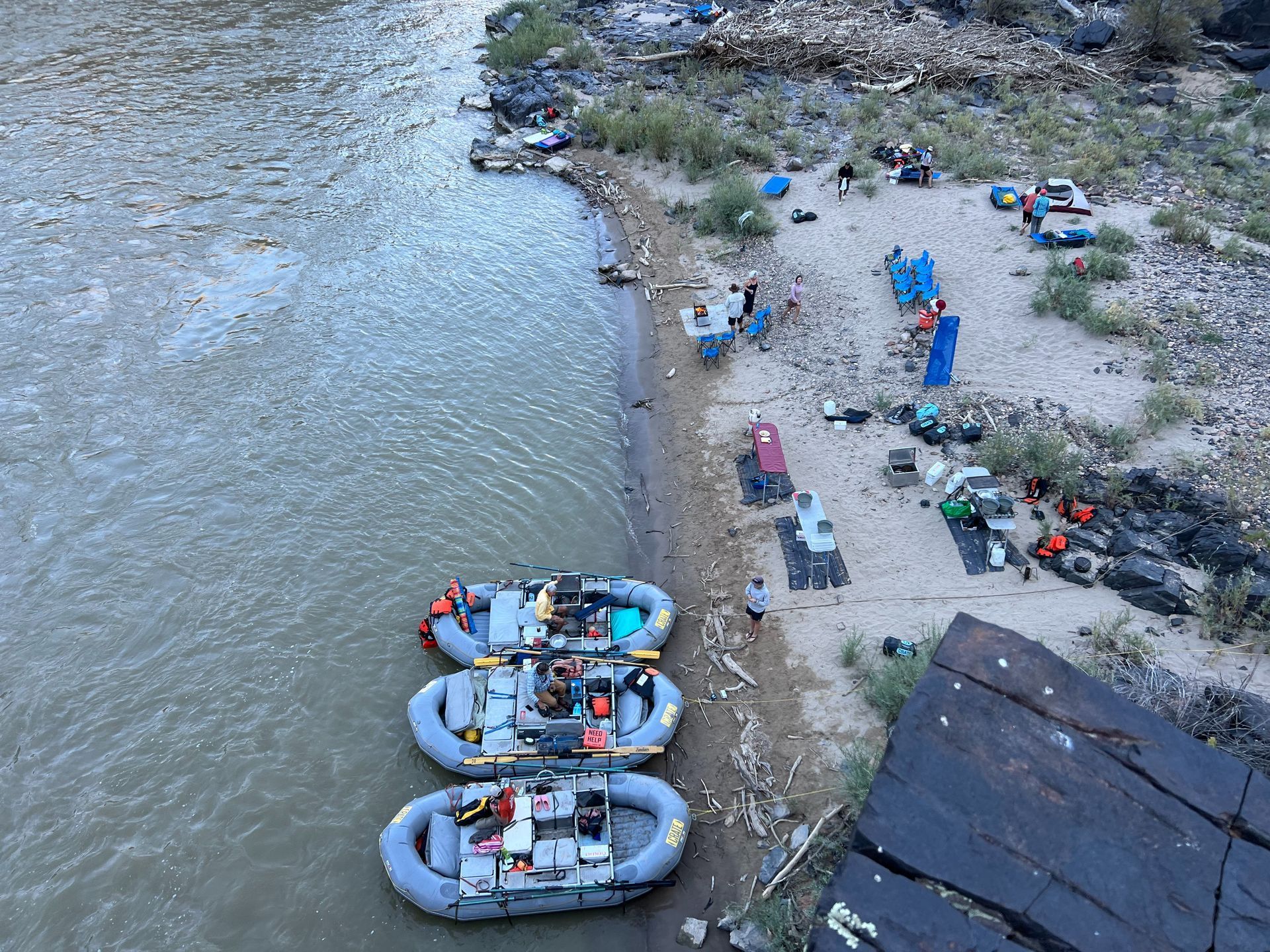 Three rafts on riverbank, people camping, unloading gear, near water and rocks.