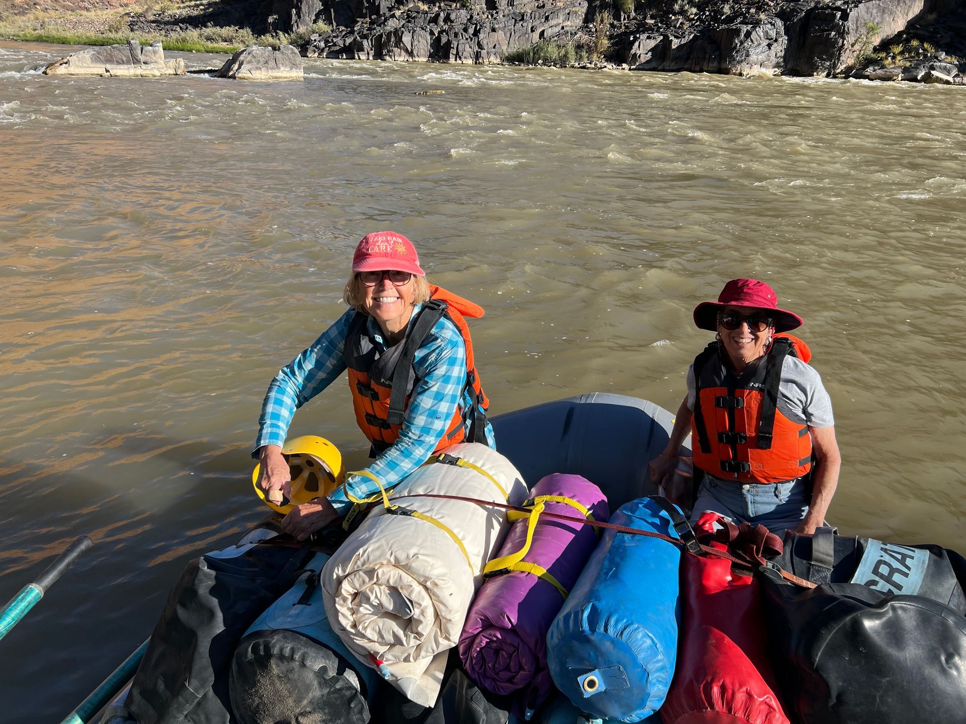 Two people in life vests on a raft, gear secured with ropes, on a river.