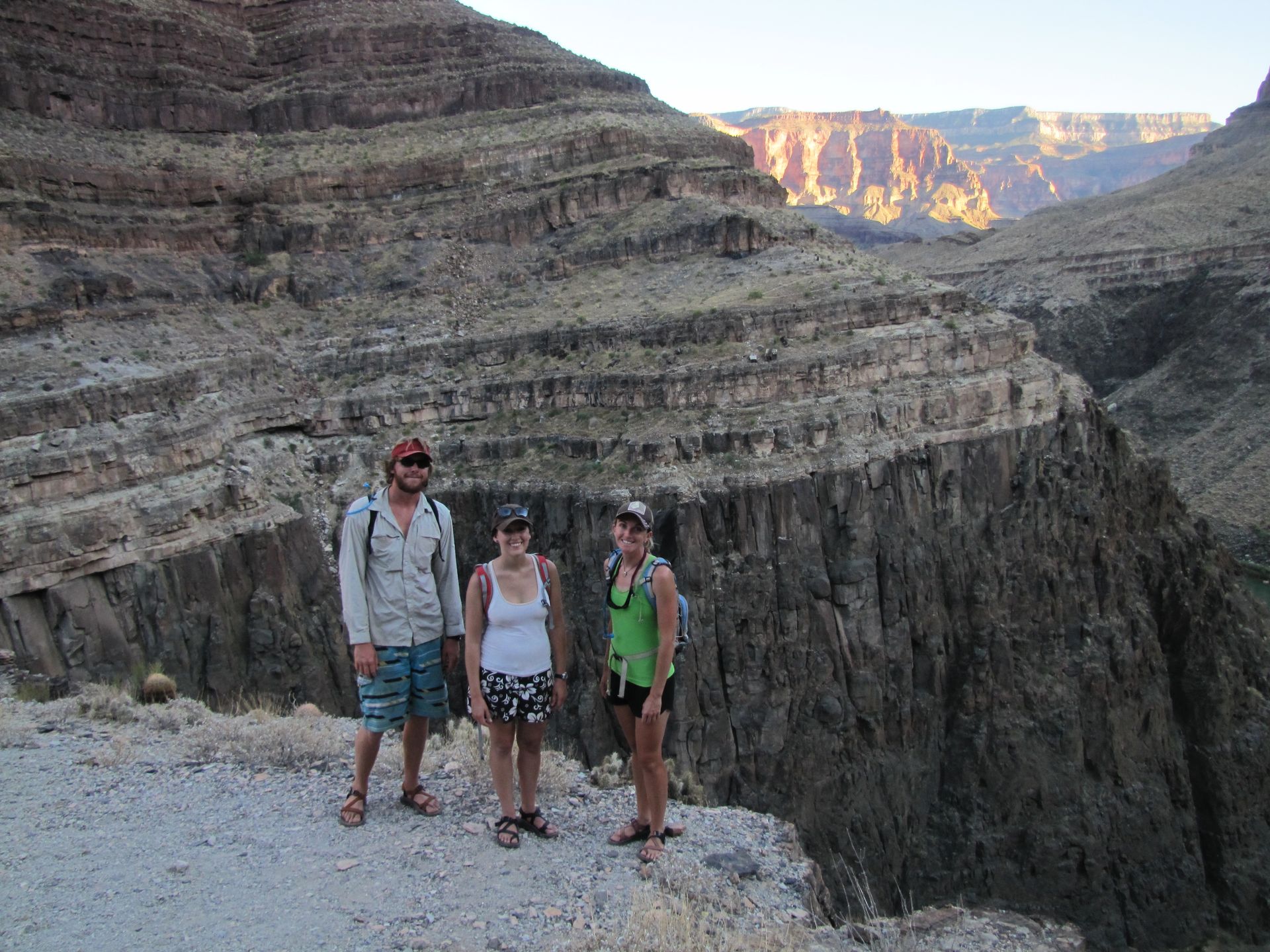 Three people stand on a canyon rim. The landscape is rocky and brown, with sunlight illuminating the distant walls.