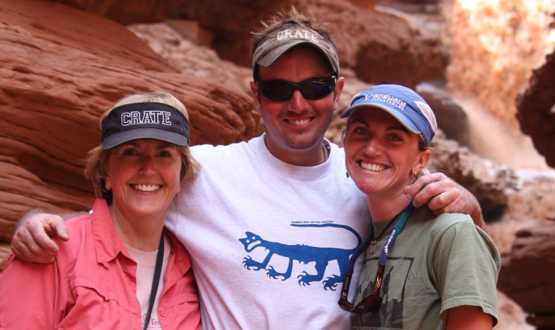 Three people smiling in a canyon. Woman on the left wears a pink shirt, man in the center wears a white shirt.