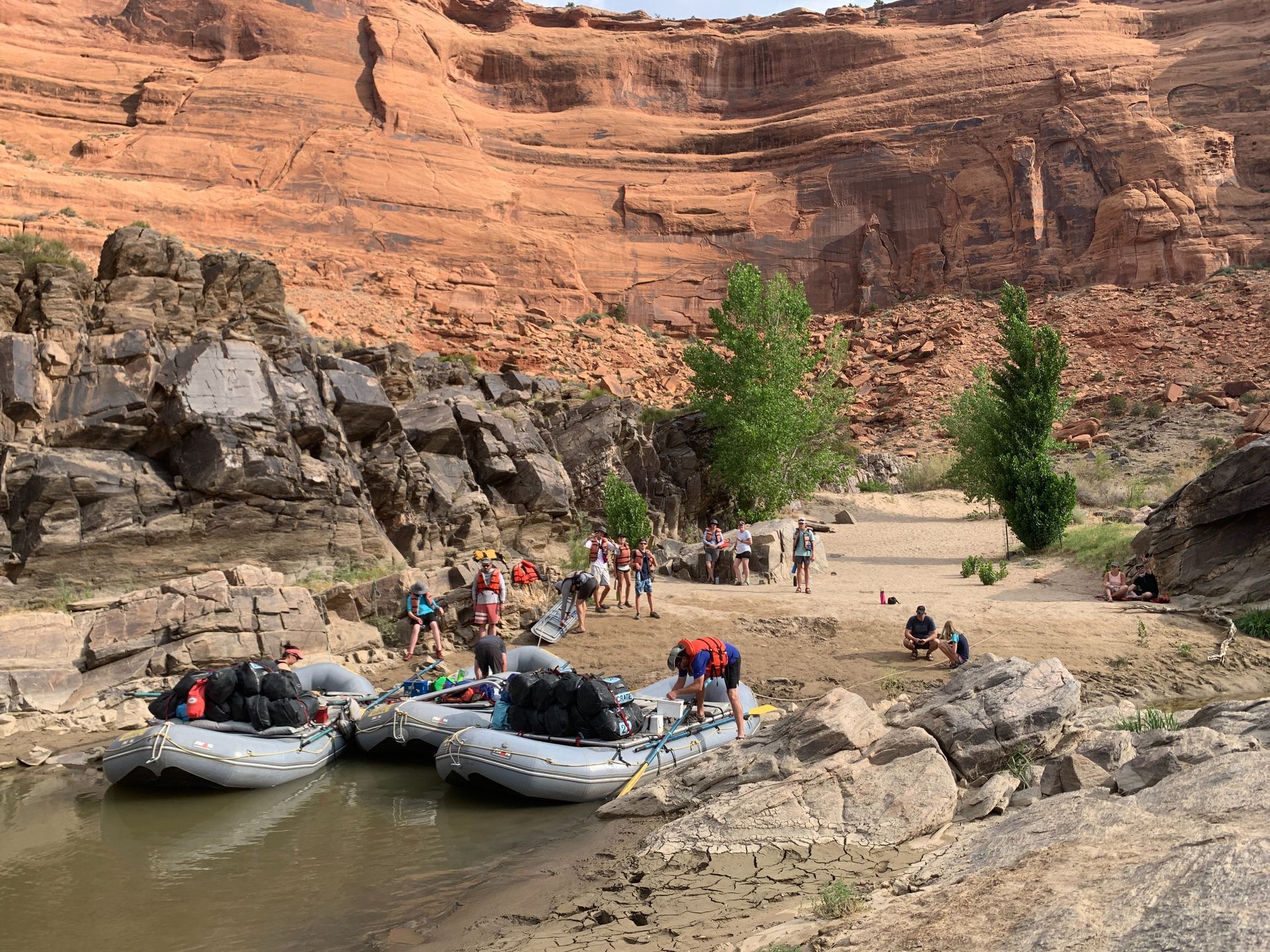 Whitewater rafts on a riverbank with people and gear. Red rock cliffs in the background.