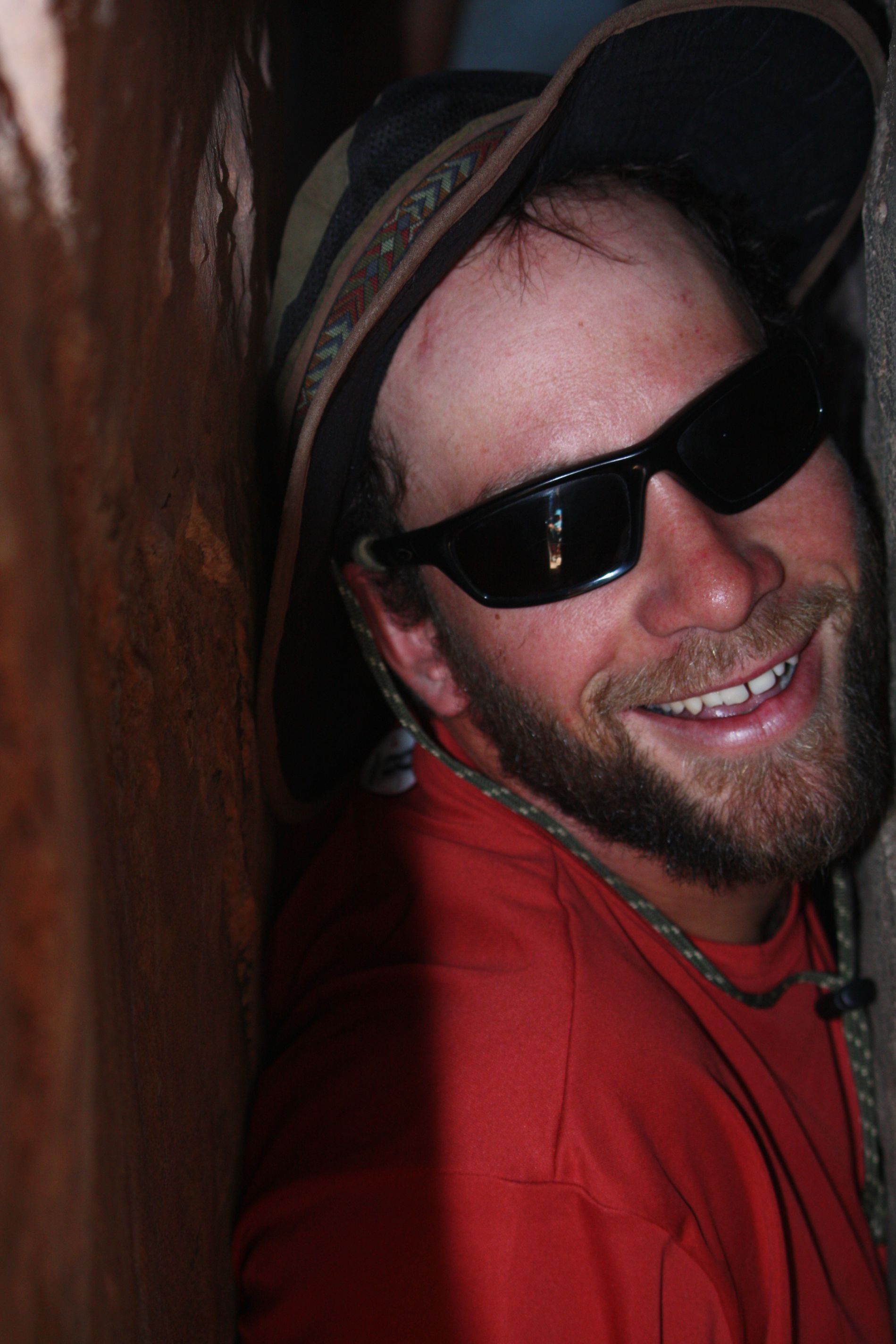 Man wearing sunglasses and a hat smiles, leaning against a brown wall, wearing an orange shirt.