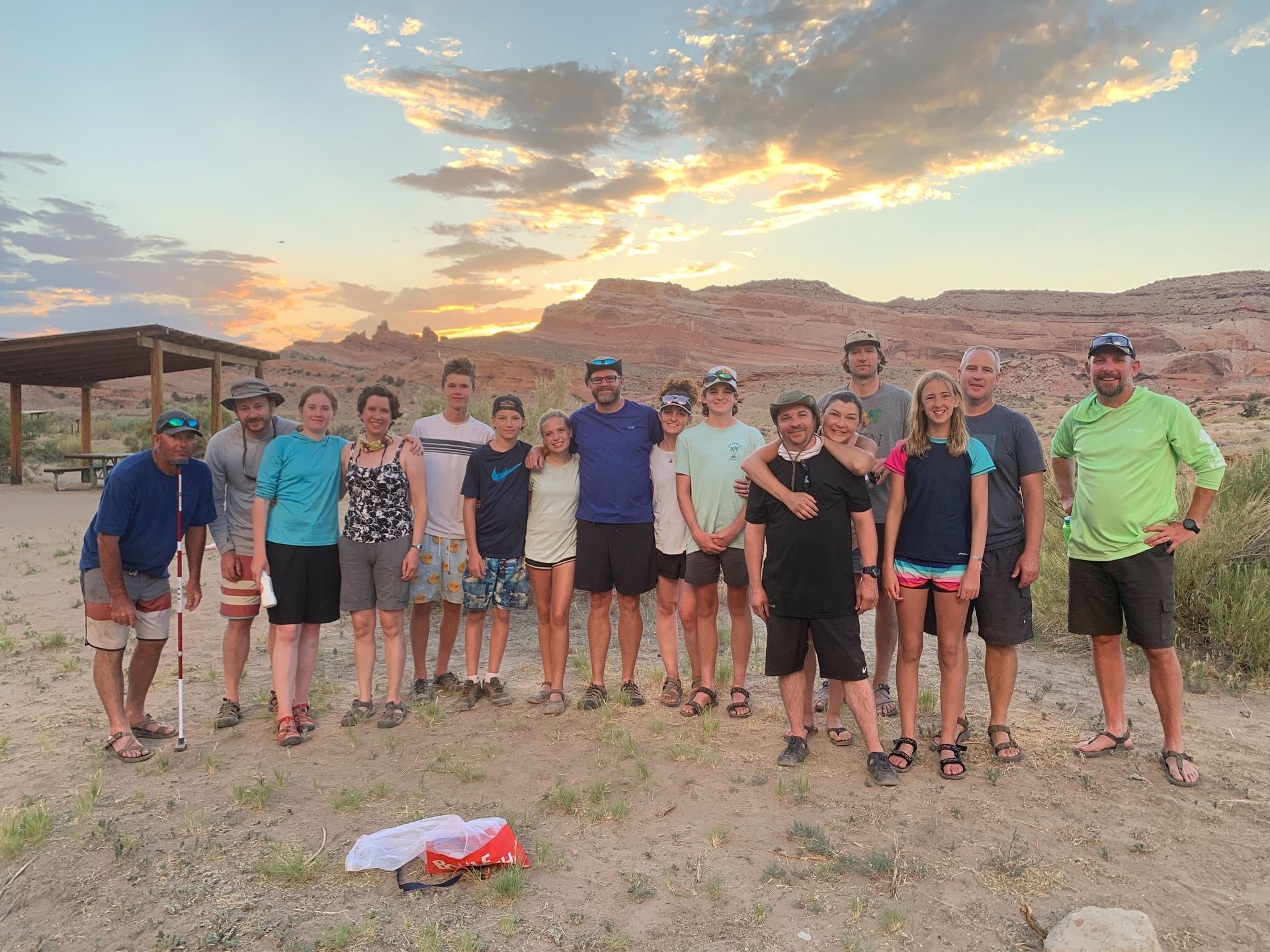 Group of people standing outdoors, posed near a shelter in front of a colorful sunset, desert landscape.