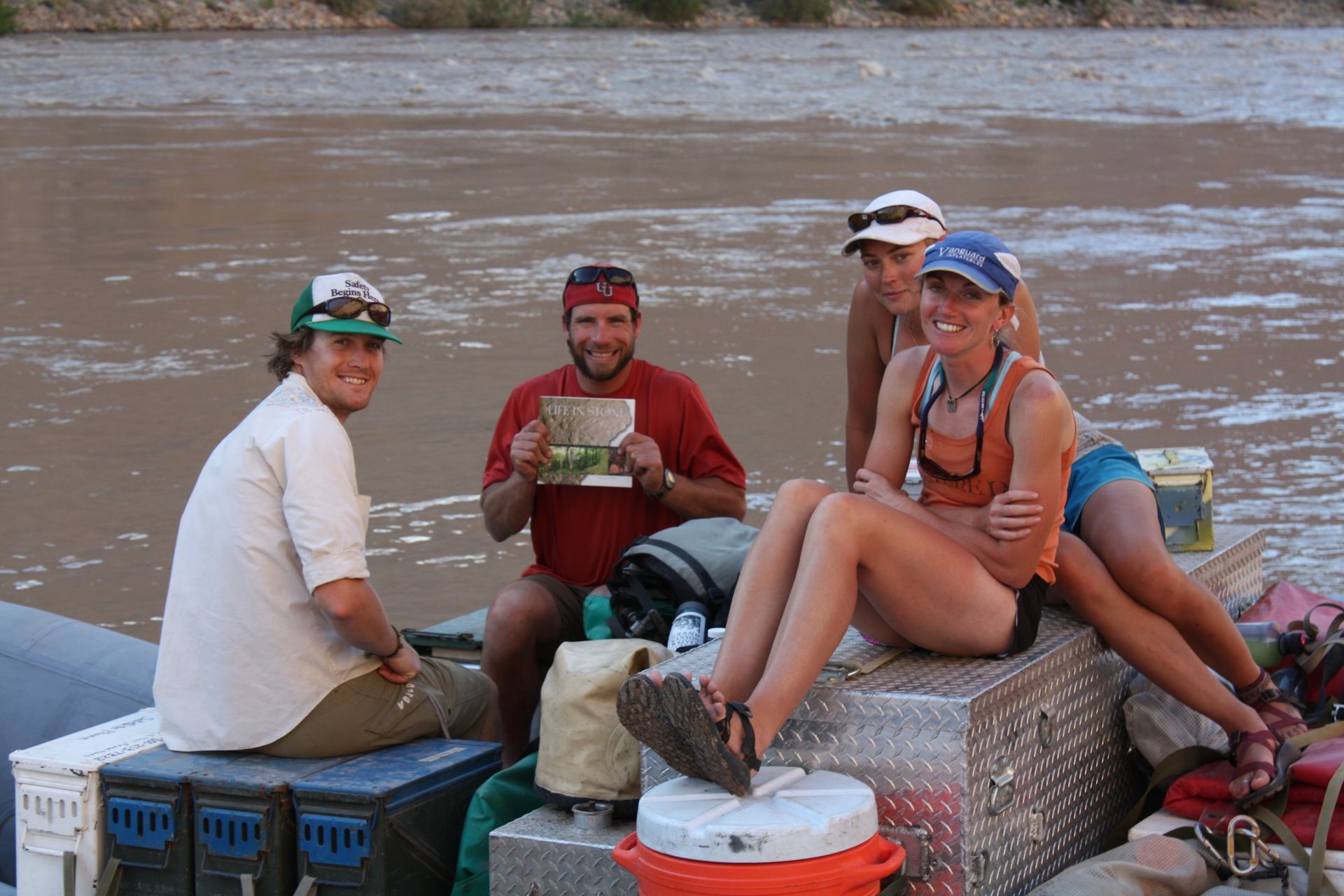 Four people on a raft on a river. One holds a drawing, all are smiling. Overcast sky, brown water.