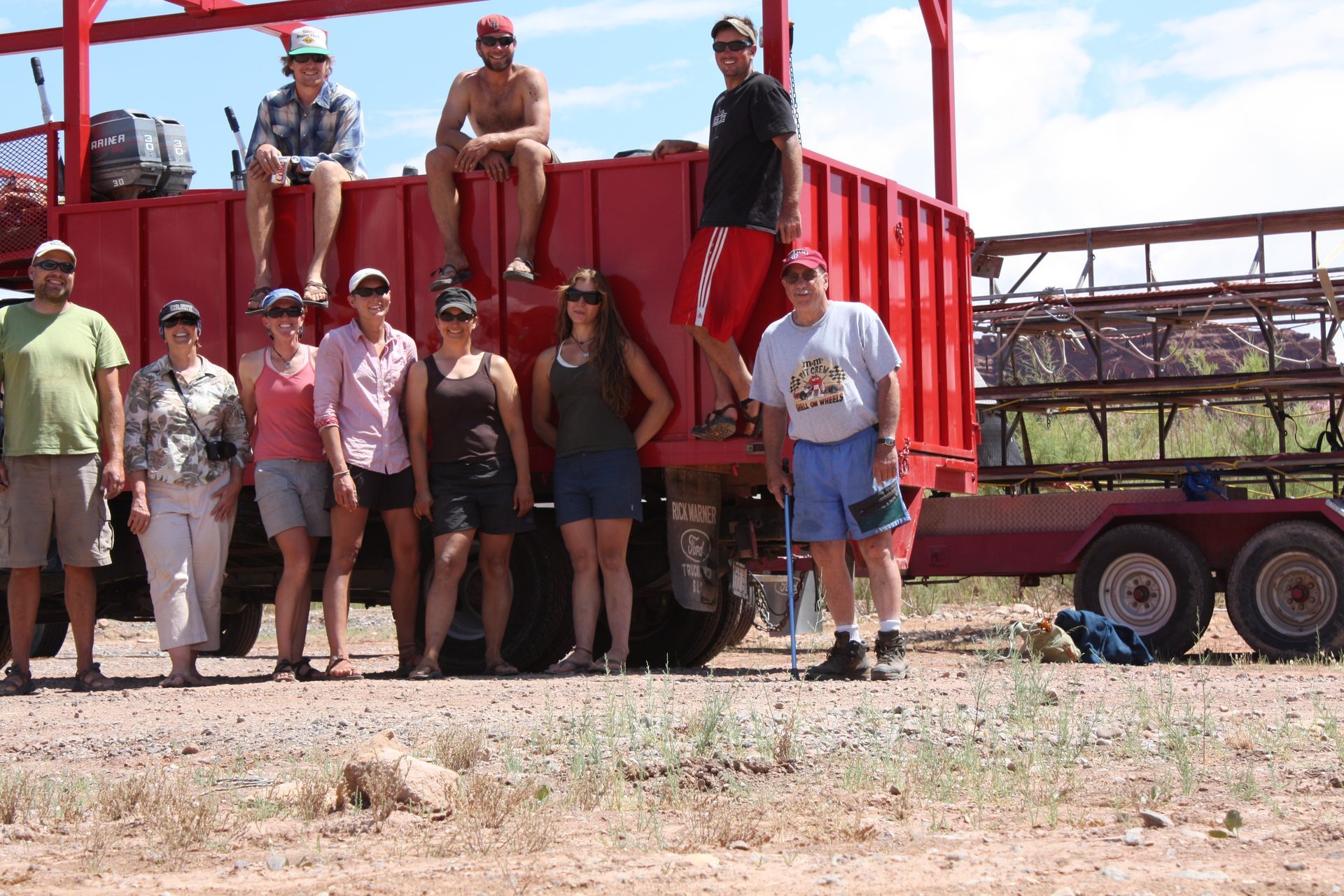 Group of people posing by a red truck trailer. Some sit on top, others stand in front on dirt. Sunny outdoor setting.