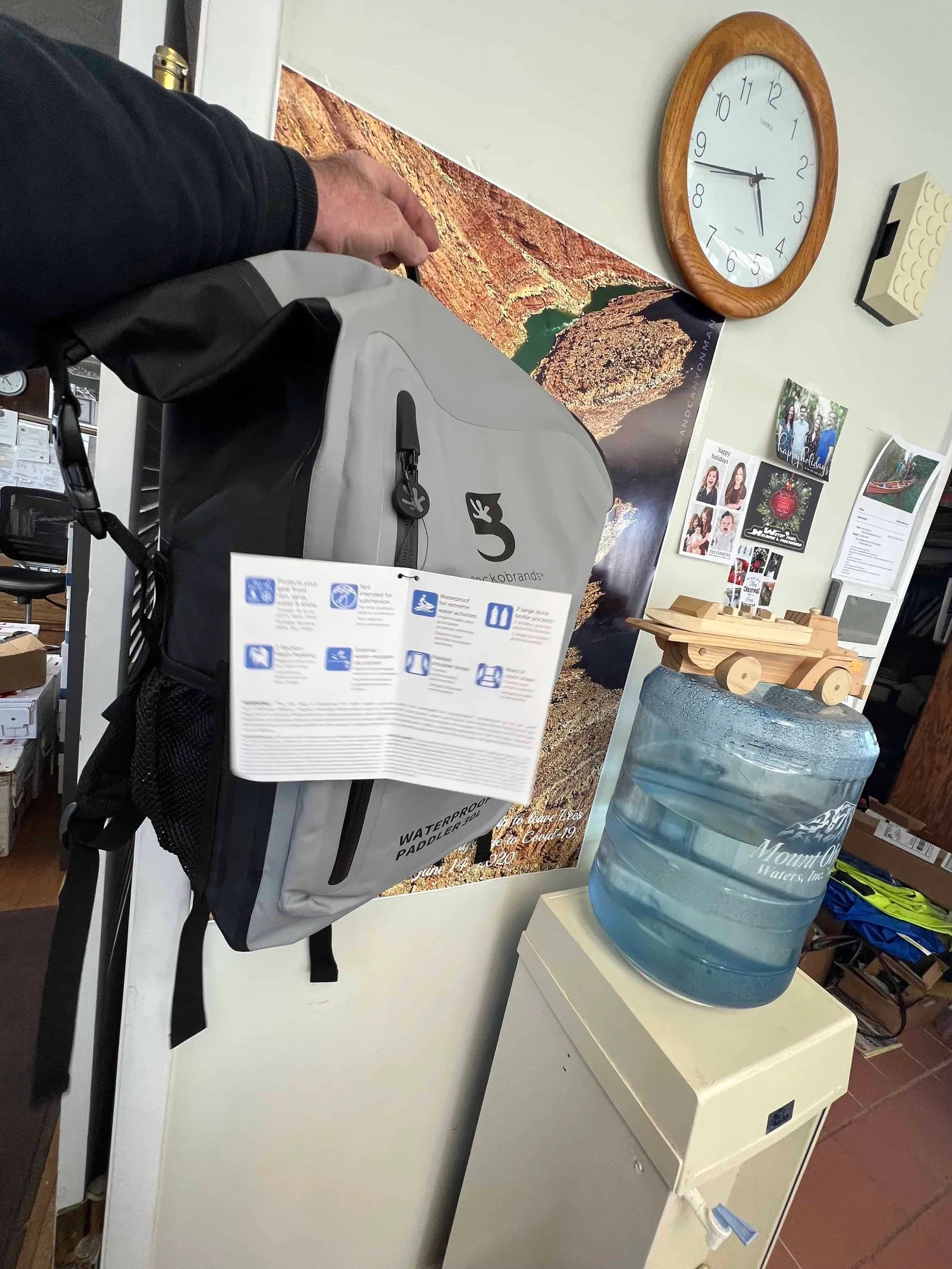 A person holding a grey and black backpack near a water cooler and a wall with a clock.