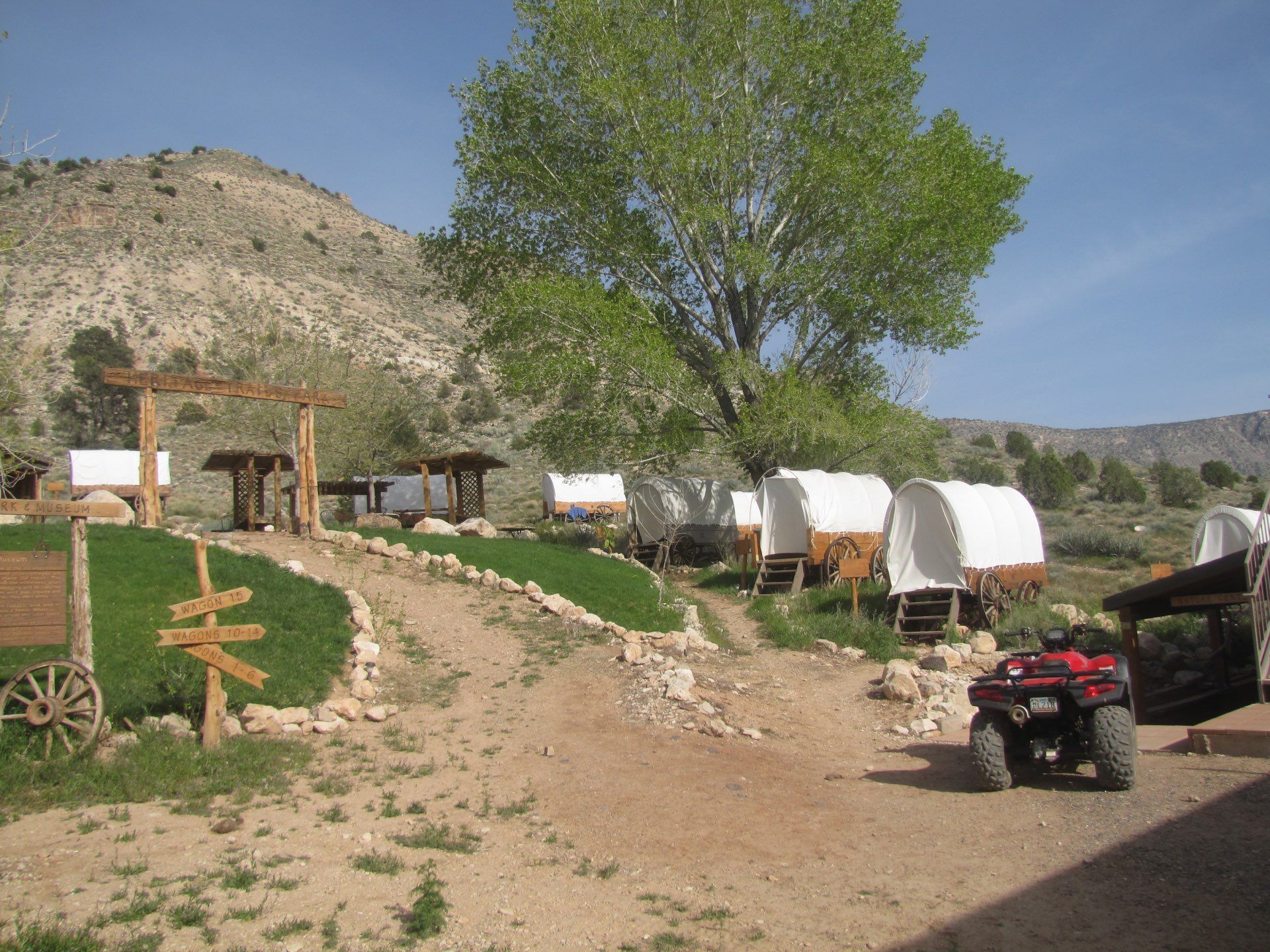 Campground with covered wagons, a wooden pergola, an ATV, and a dirt path leading toward a mountain.