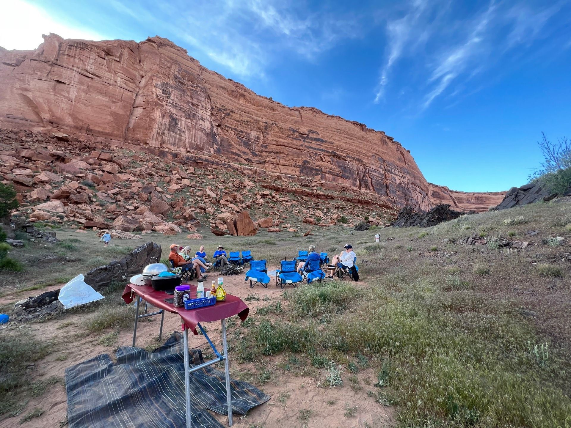 Campers gathered by a red rock cliff. Blue chairs, table with supplies, and blue sky.
