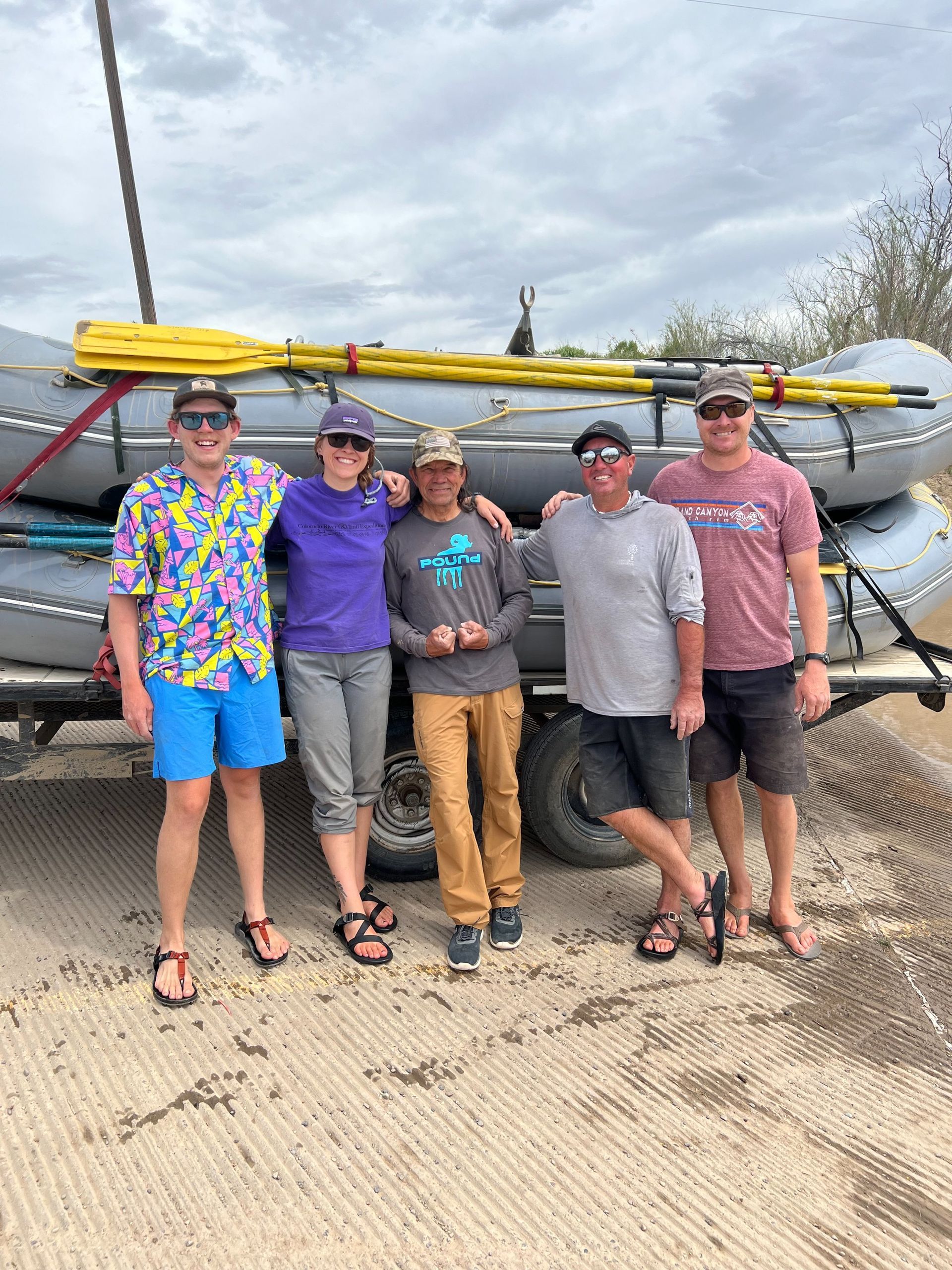 Five people stand in front of a raft on a trailer, posing. Cloudy sky backdrop.