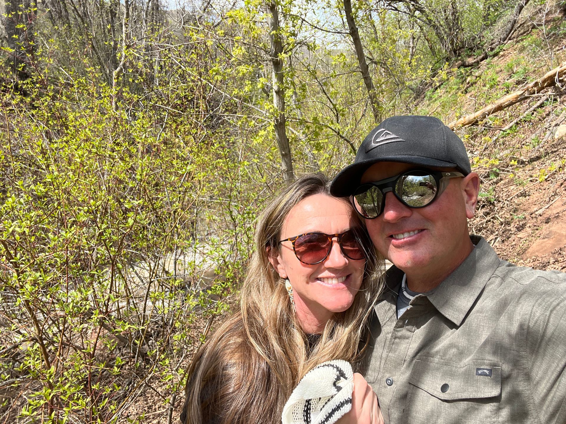 Couple smiling on a sunny hiking trail, surrounded by green foliage.