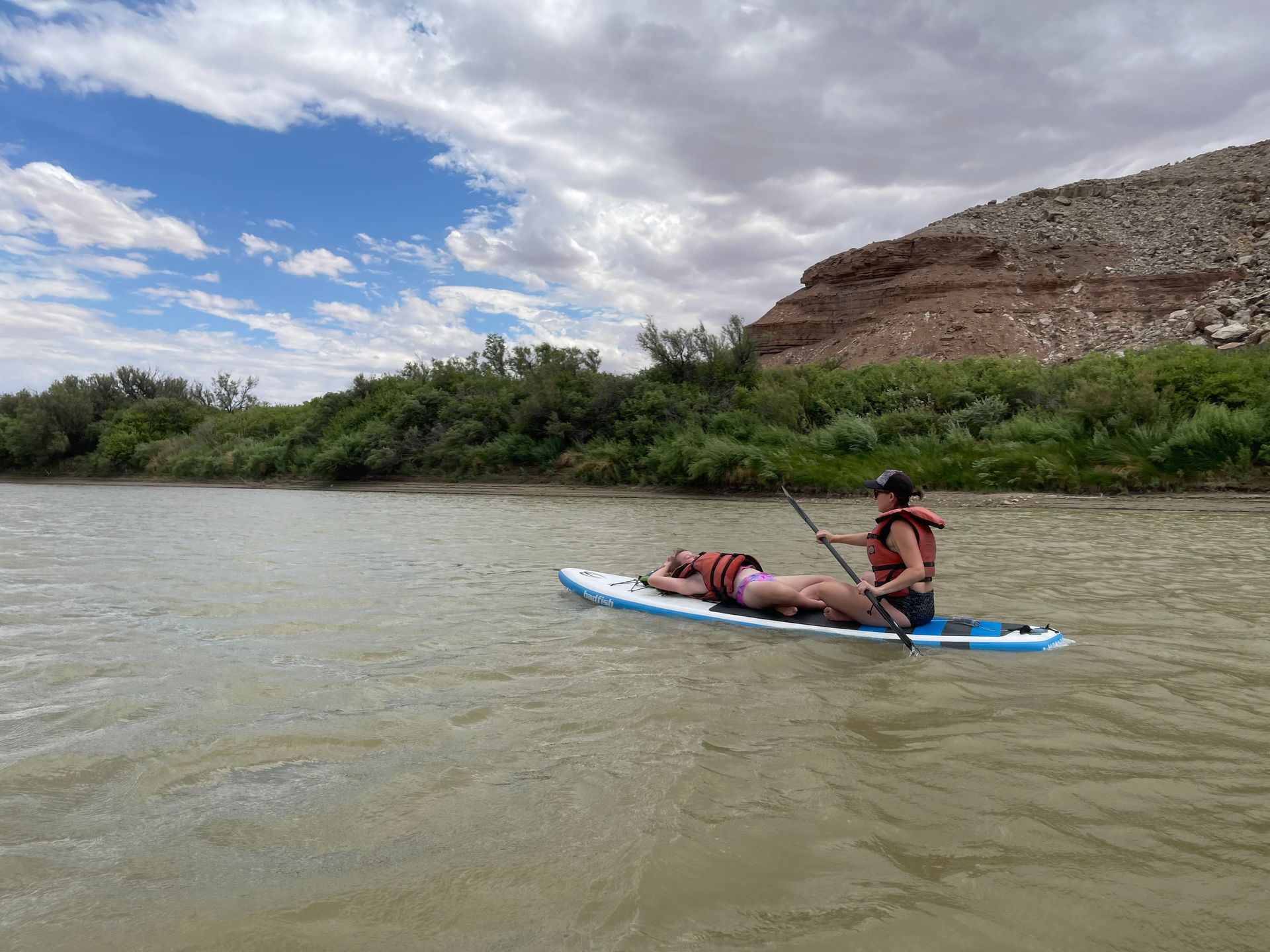 Two people paddleboarding on a river near a rocky hillside. One lies down. Cloudy sky.