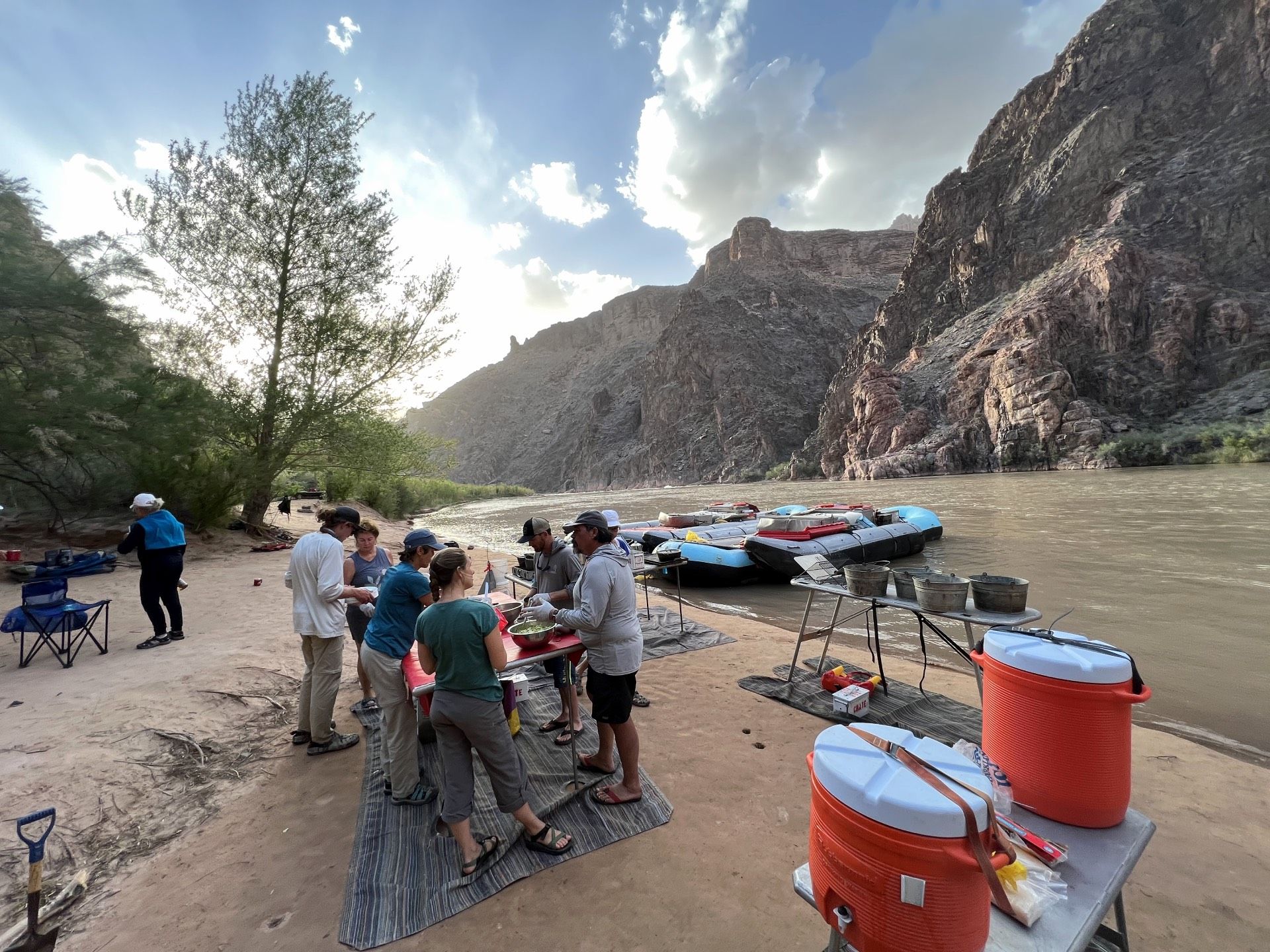 People preparing food at a riverside campsite with rafts. Mountains and sky in the background.