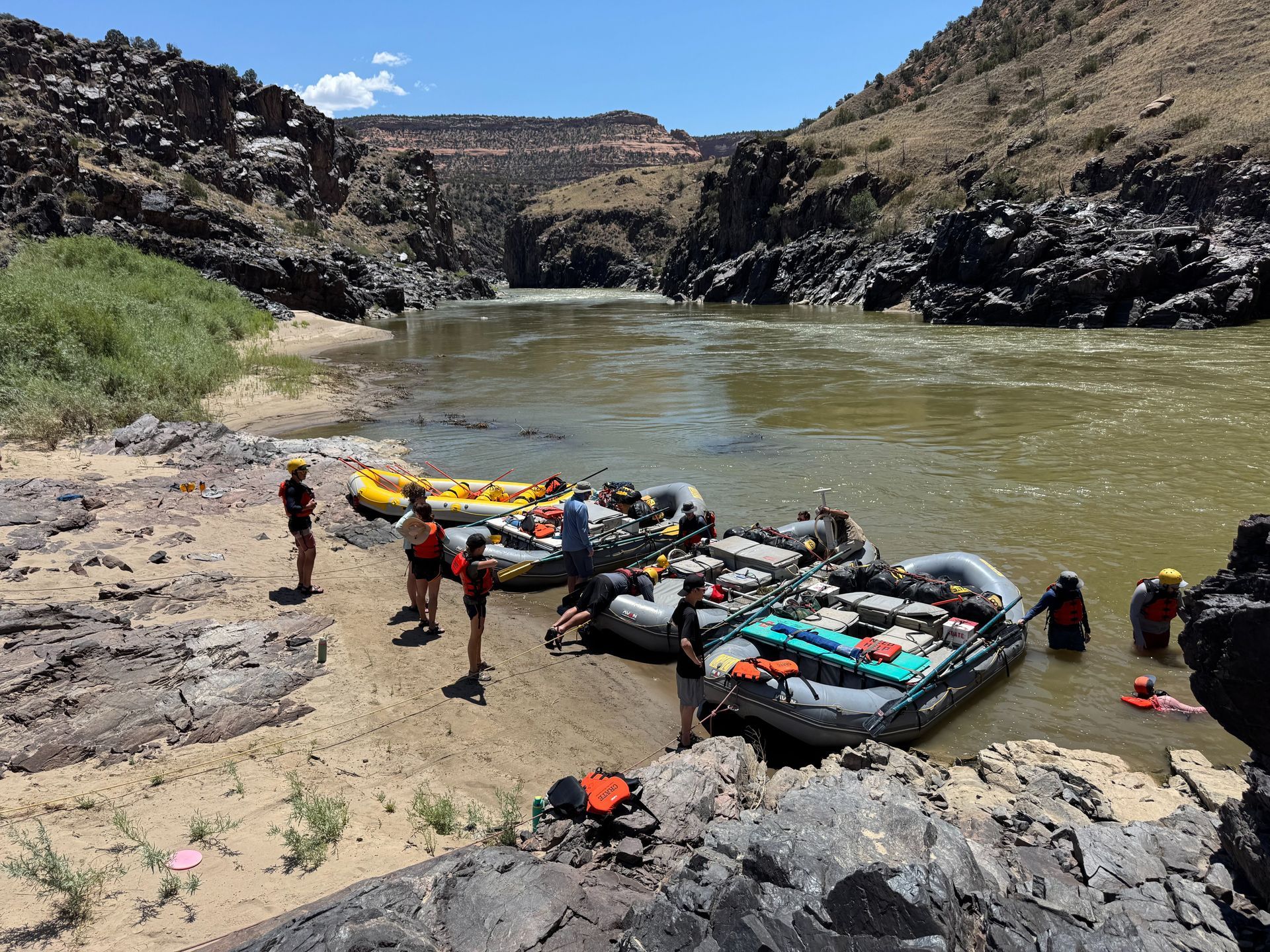 People and rafts on a riverbank near rocky cliffs.