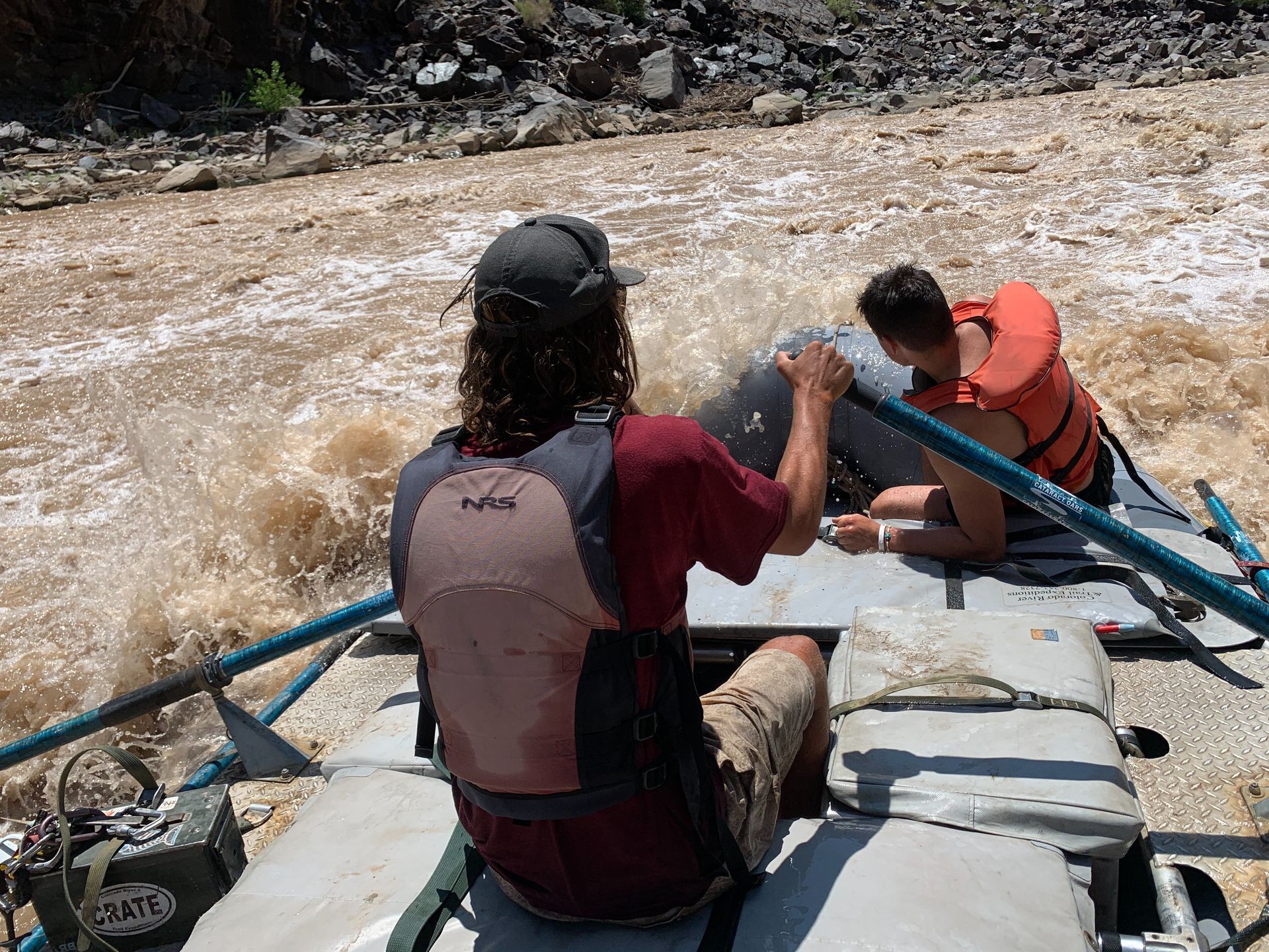 Two people on a raft navigating a turbulent, muddy river. One rows, other holds on.